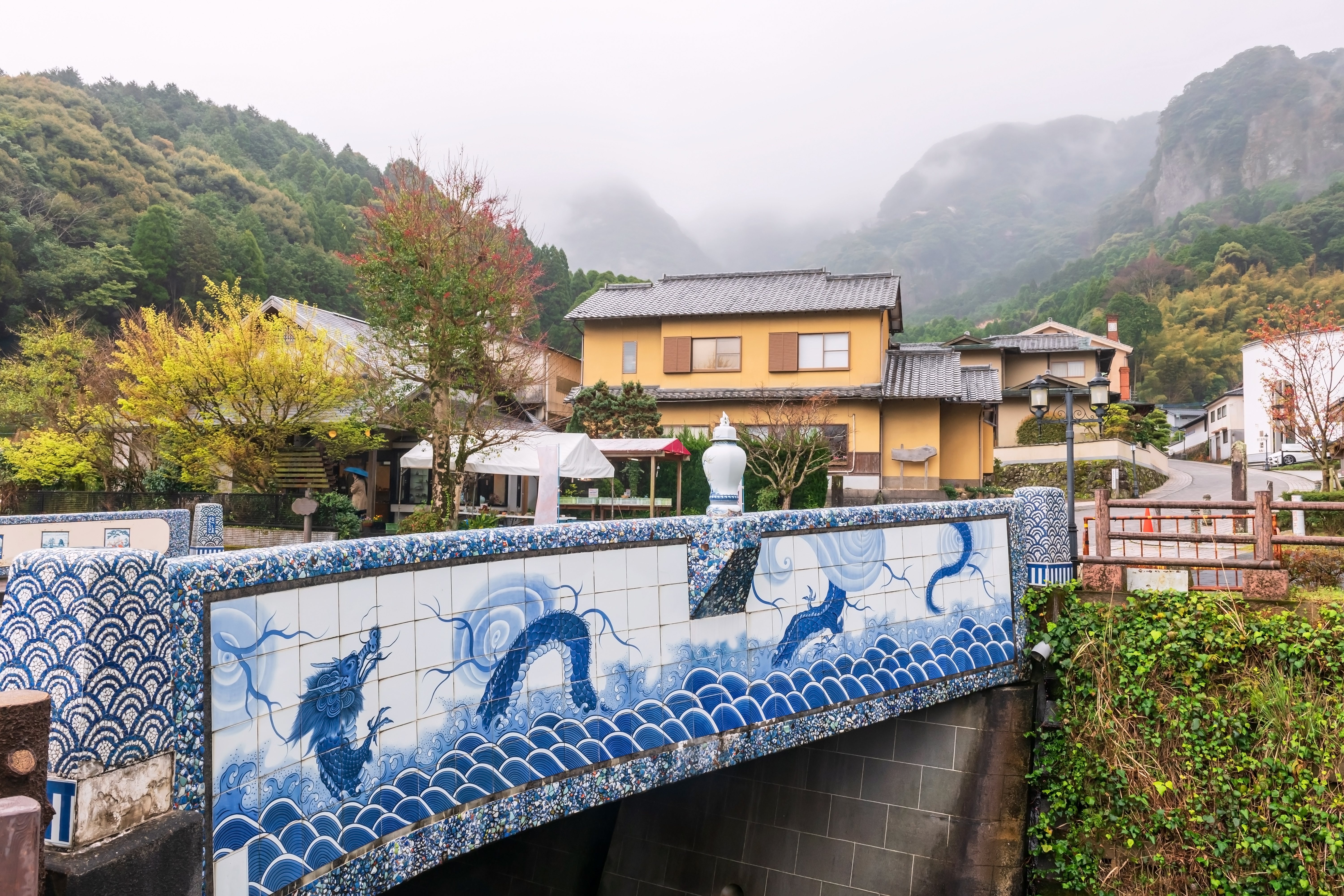 A porcelain-decorated bridge spans a narrow canal in Okawachiyama village, Saga, with misty mountains behind.