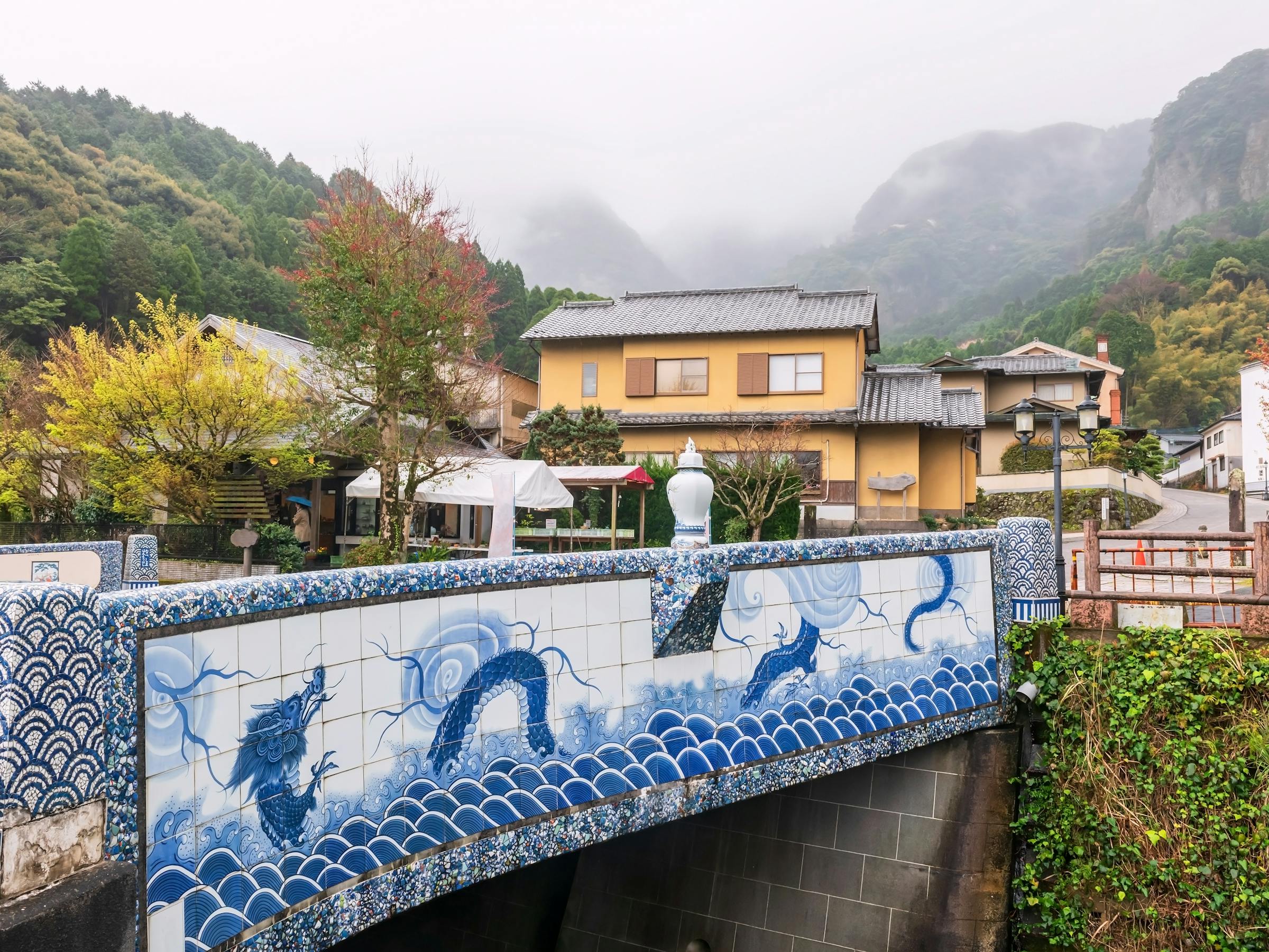 A porcelain-decorated bridge spans a narrow canal in Okawachiyama village, Saga, with misty mountains behind.