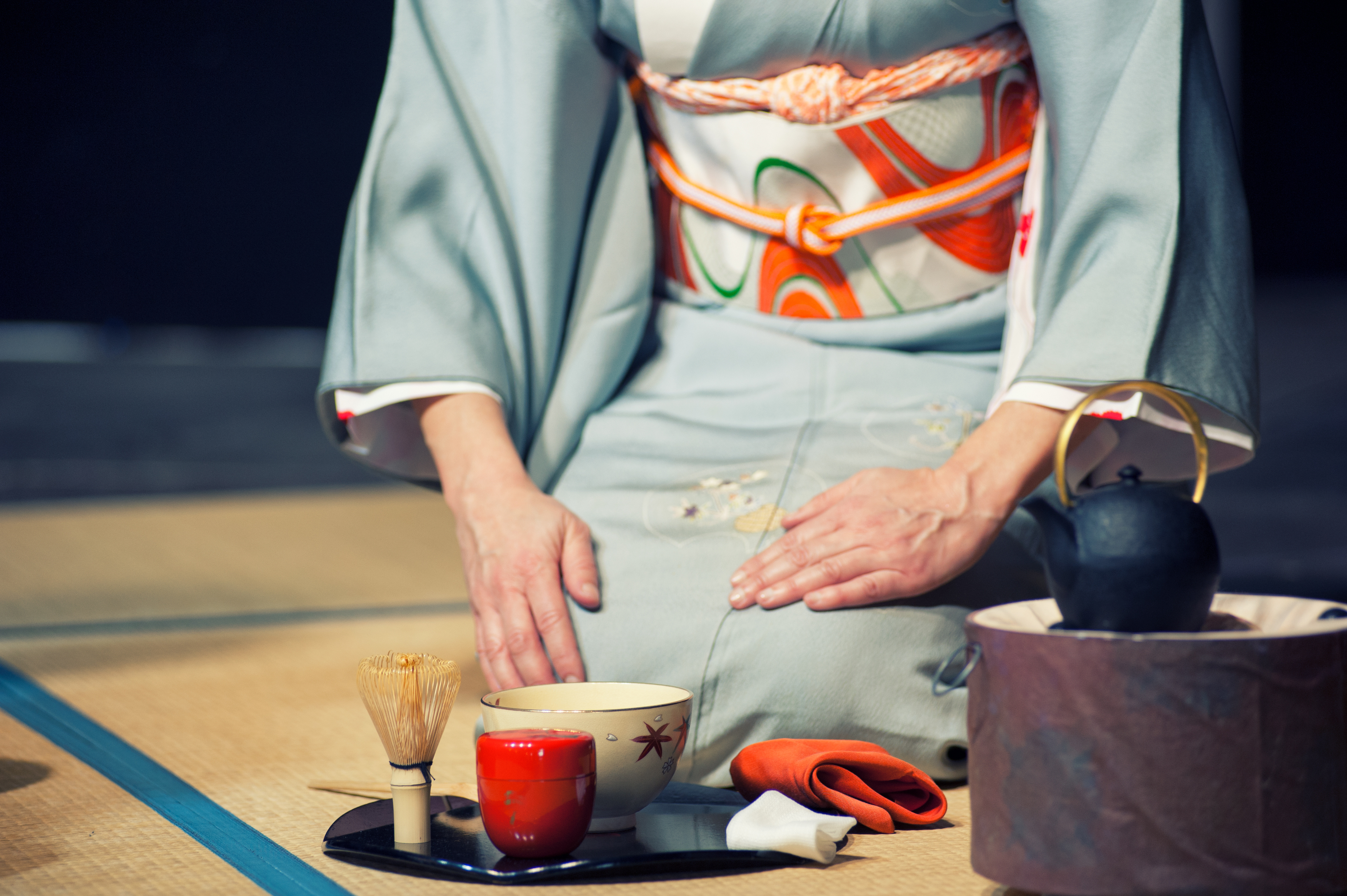 A woman in a pale kimono presents a teacup during a tea ceremony, with utensils and a kettle set beside her.