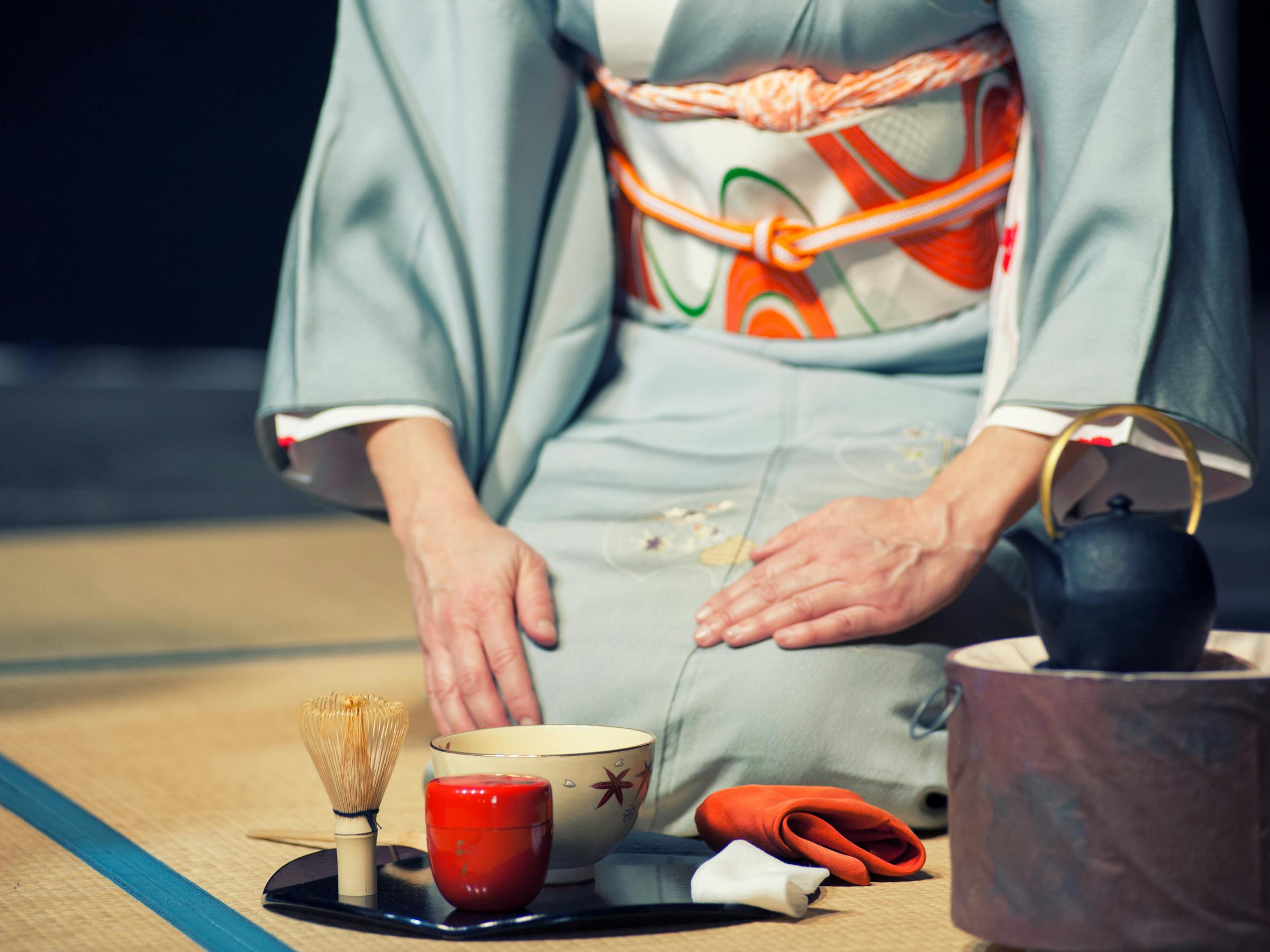 A woman in a pale kimono presents a teacup during a tea ceremony, with utensils and a kettle set beside her.