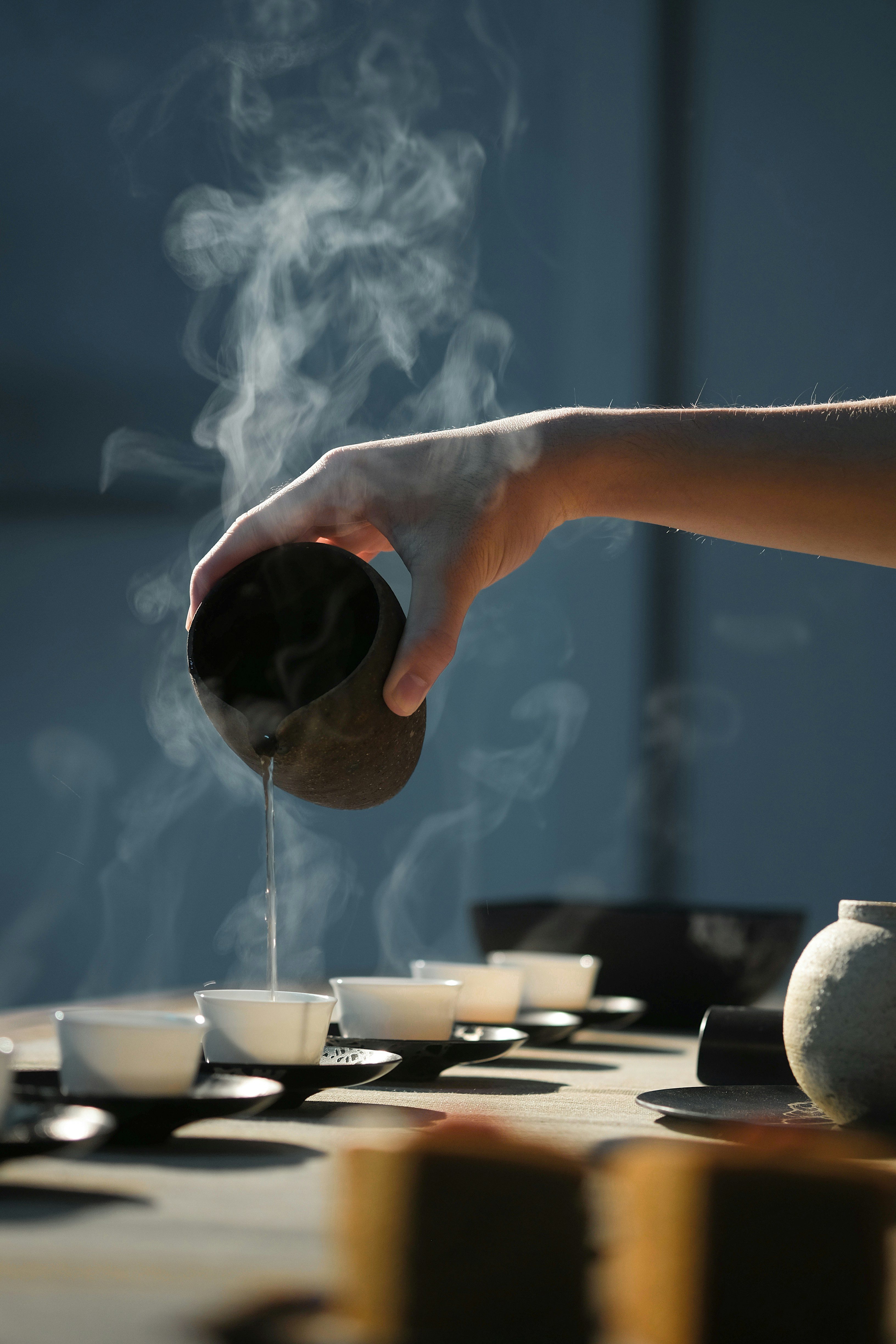 A hand pours hot tea into small cups as steam curls upward, with a tea tray and bowls softly lit on a table.