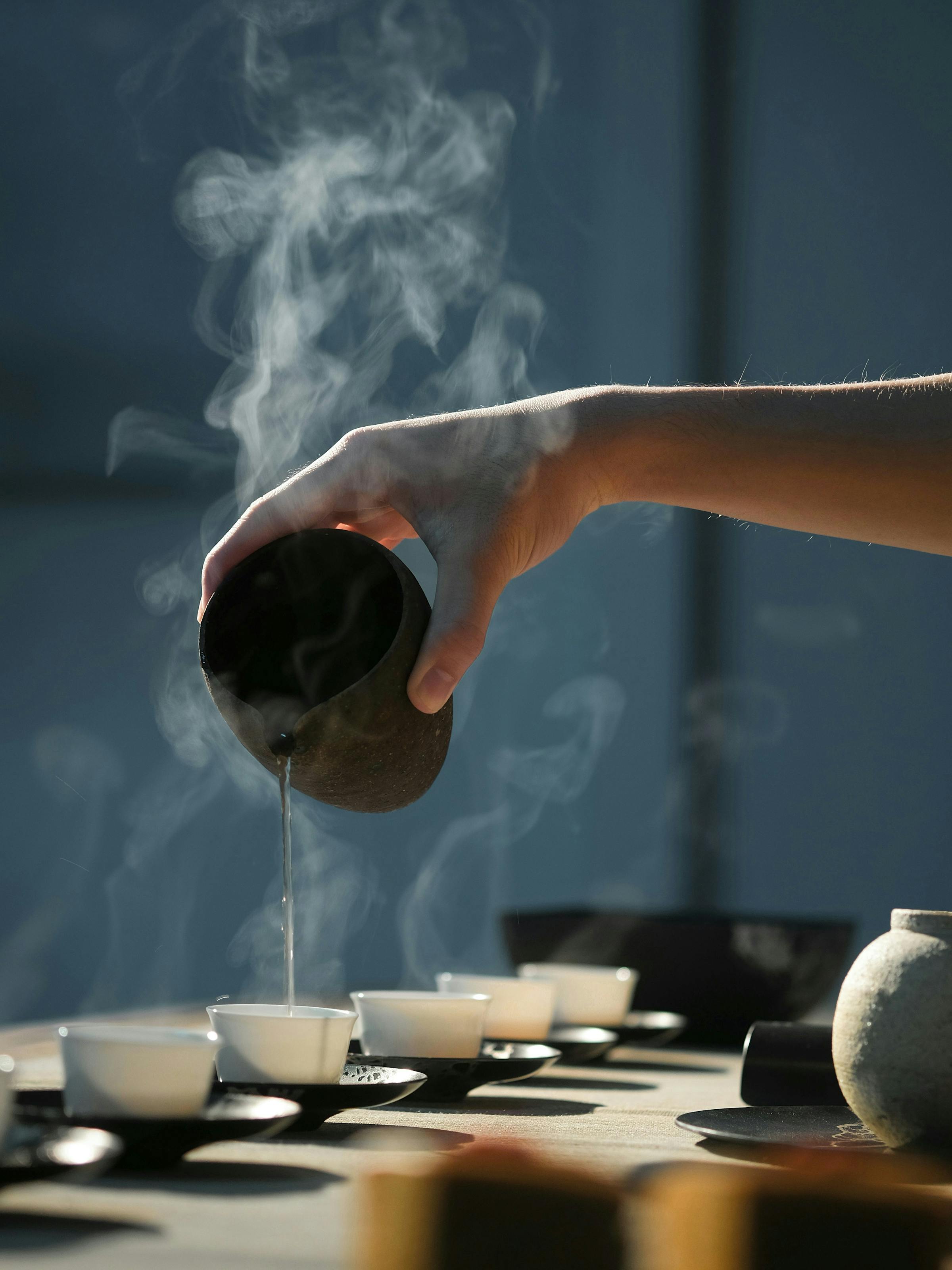 A hand pours hot tea into small cups as steam curls upward, with a tea tray and bowls softly lit on a table.