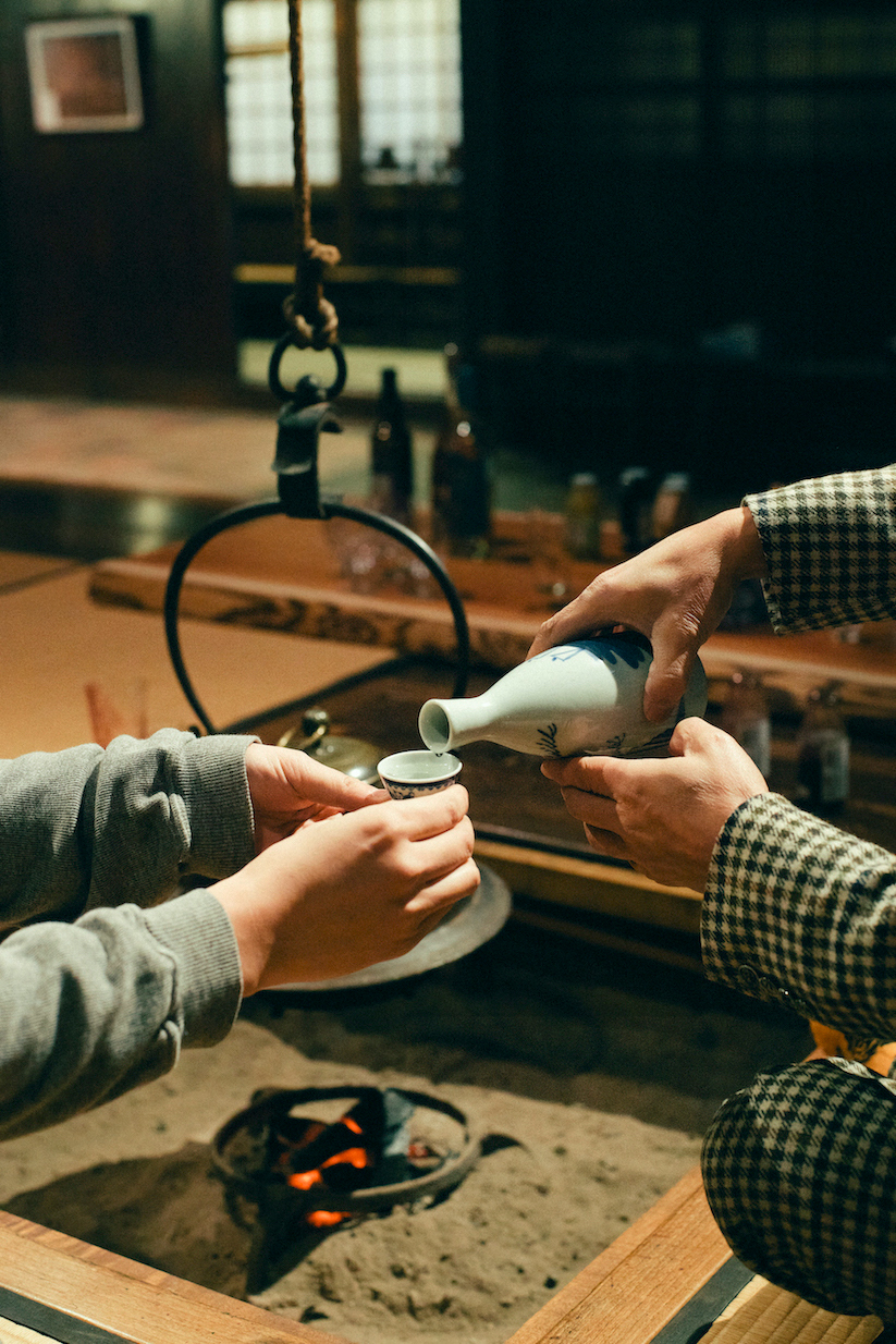 Hands pour a drink into a small cup at a low hearth table, with warm lantern light and a kettle behind in soft light.