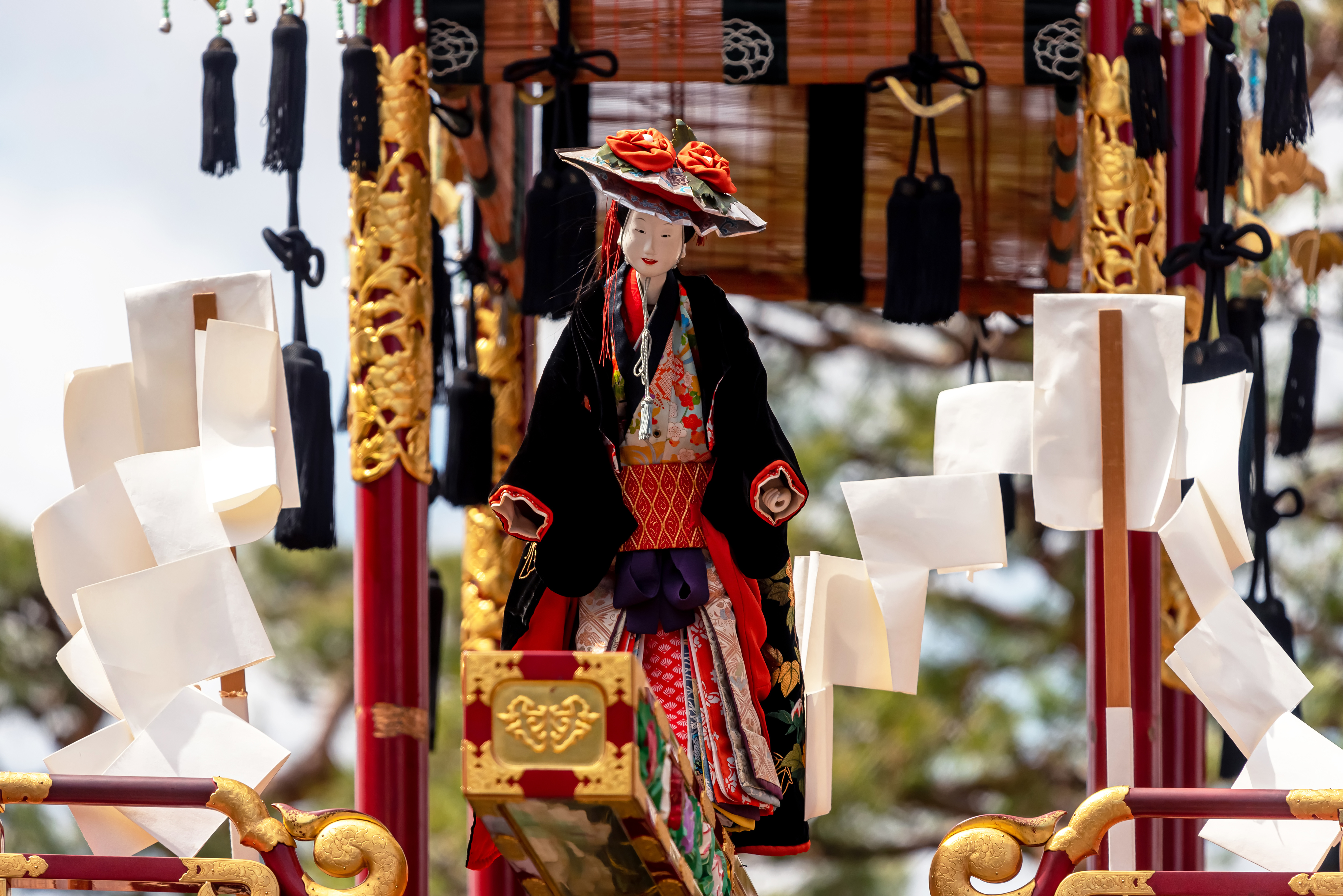 A richly dressed puppet sits on an ornate Takayama festival float, framed by carved wood and hanging tassels.