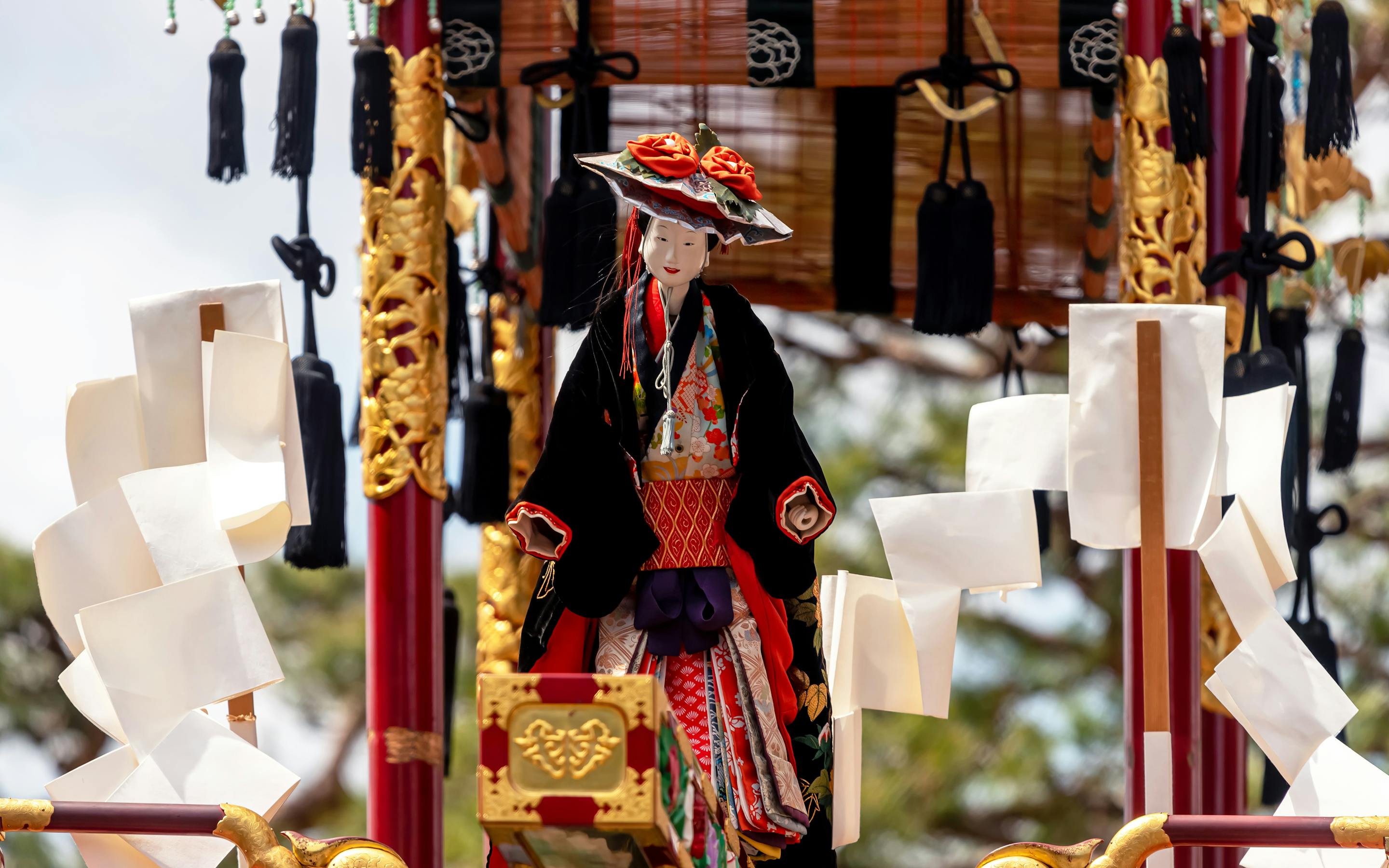 A richly dressed puppet sits on an ornate Takayama festival float, framed by carved wood and hanging tassels.