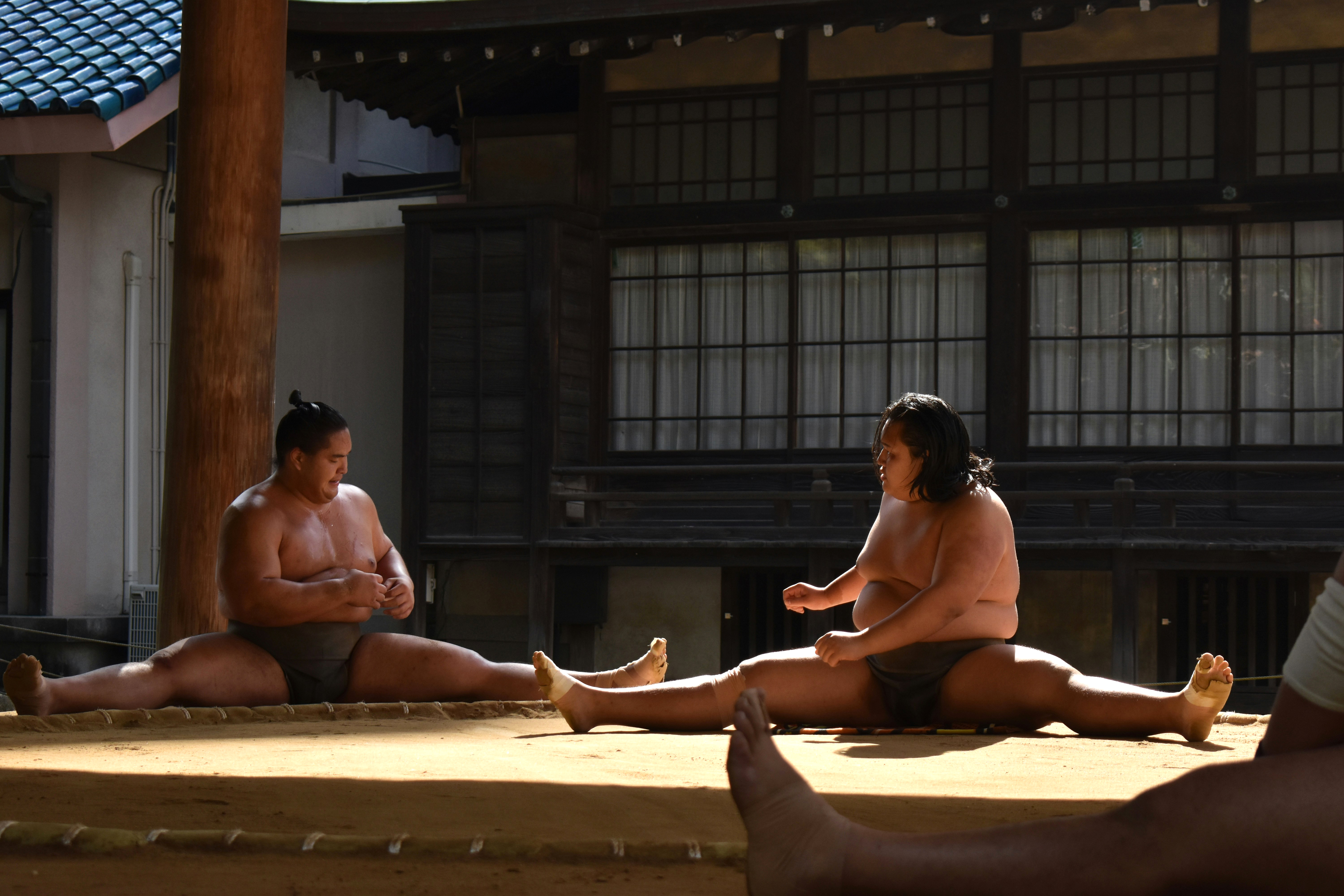 Two sumo wrestlers stretch on a raised ring in Fukuoka, with a traditional building and spectators around the dohyo.