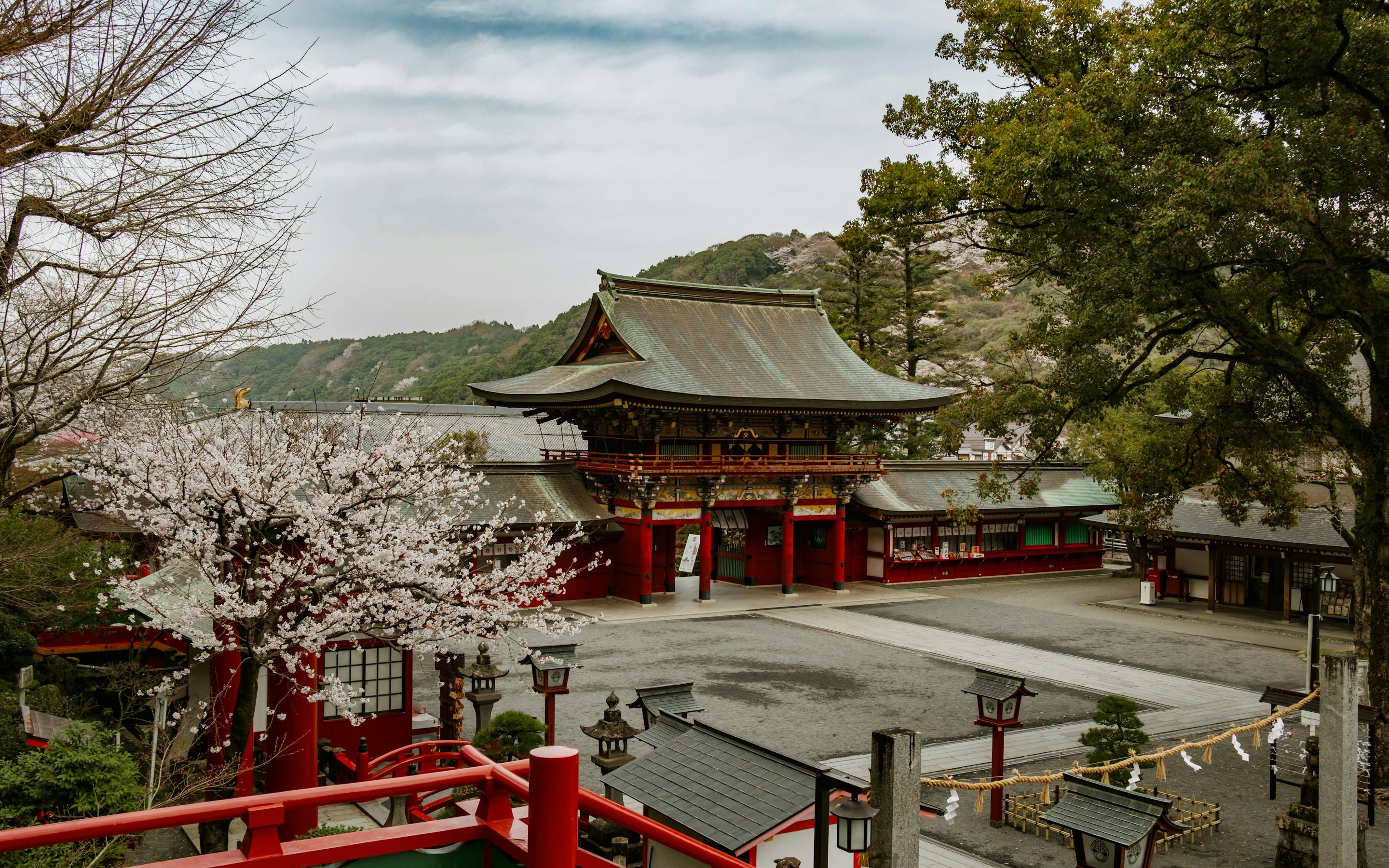 Yūtoku Inari Shrine’s red gate and stairway overlook a quiet courtyard, framed by cherry blossoms and hills.