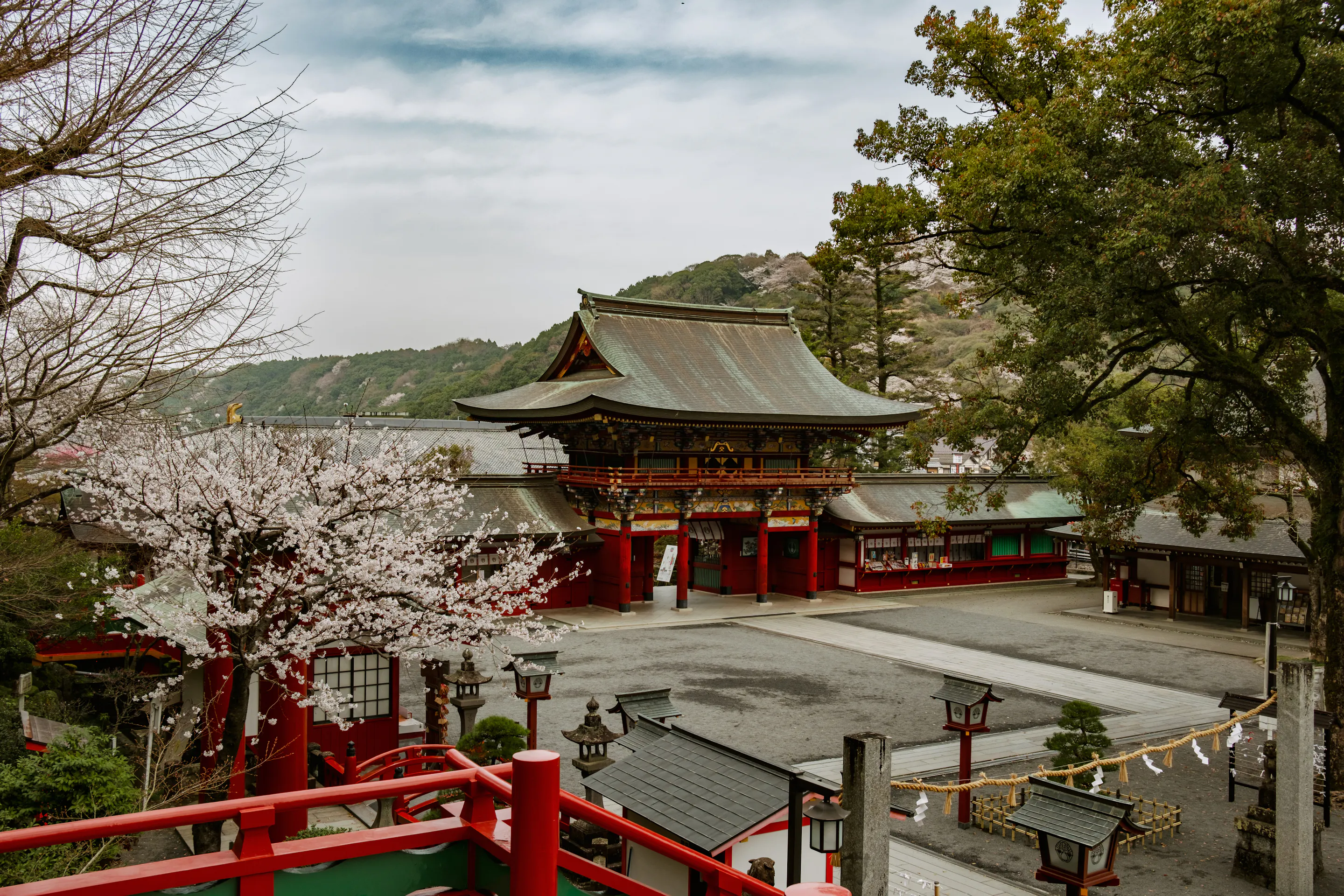 Yūtoku Inari Shrine’s red gate and stairway overlook a quiet courtyard, framed by cherry blossoms and hills.