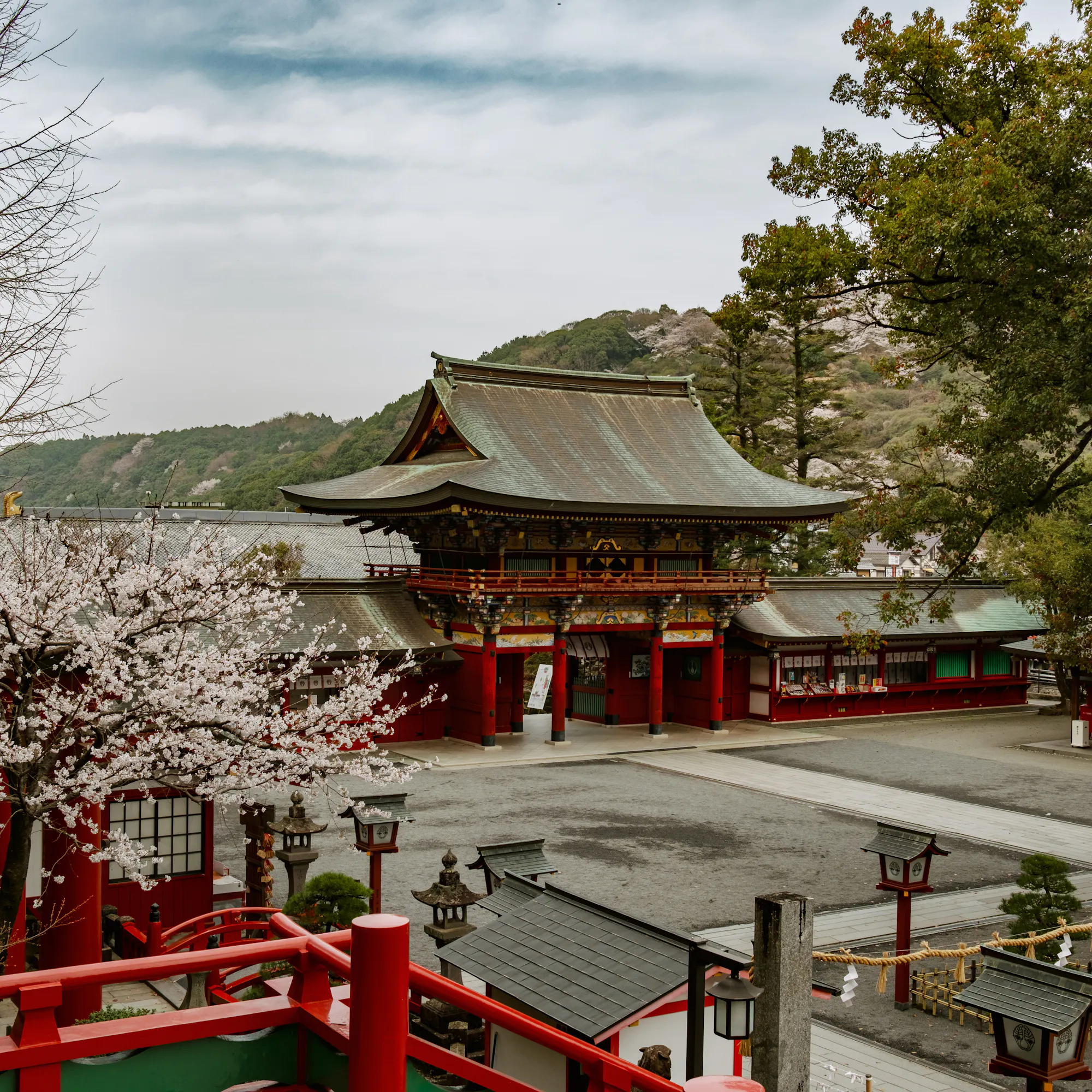 Yūtoku Inari Shrine’s red gate and stairway overlook a quiet courtyard, framed by cherry blossoms and hills.