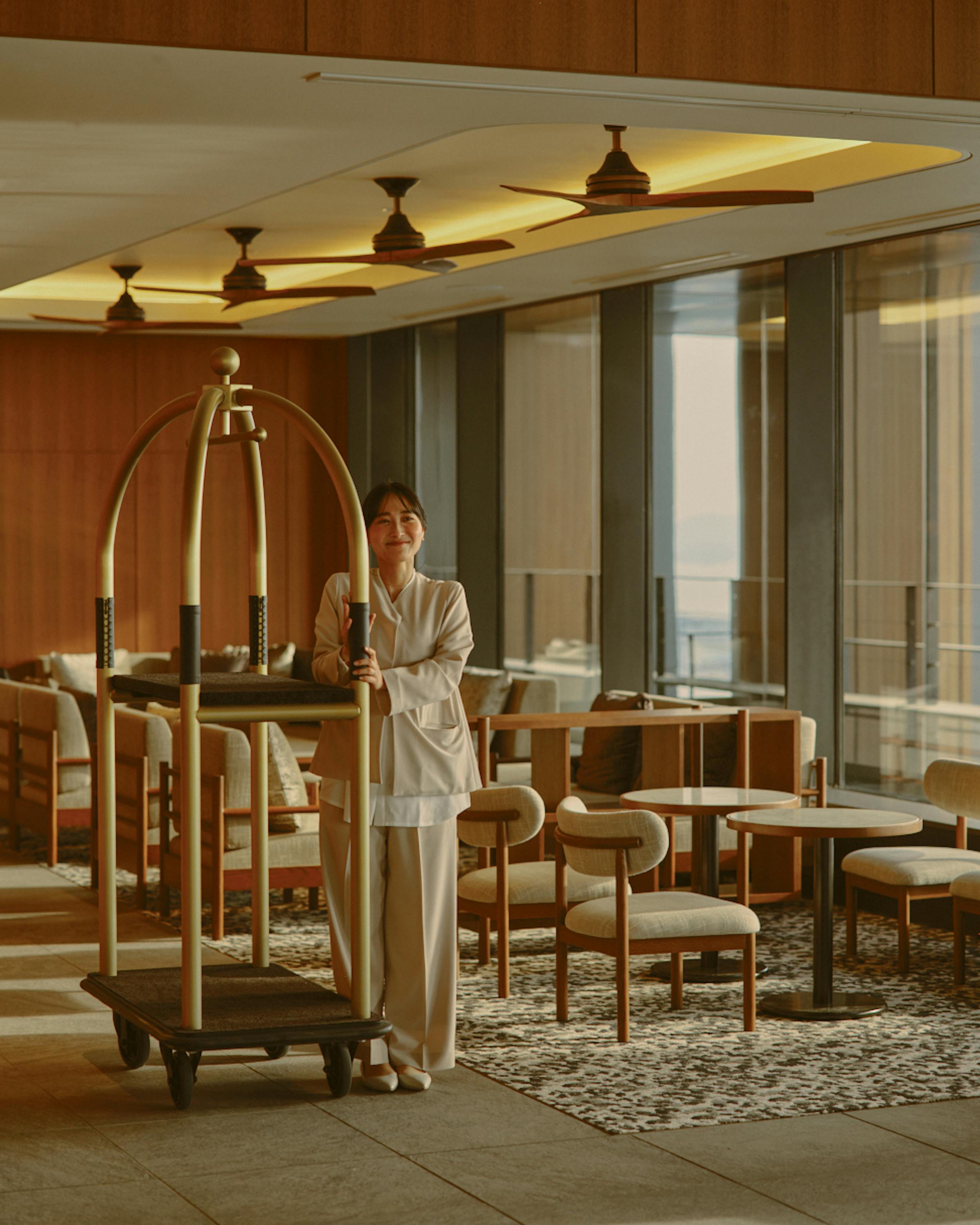 A hotel staff member stands with a luggage cart in a bright lounge, with seating and city views through tall windows.