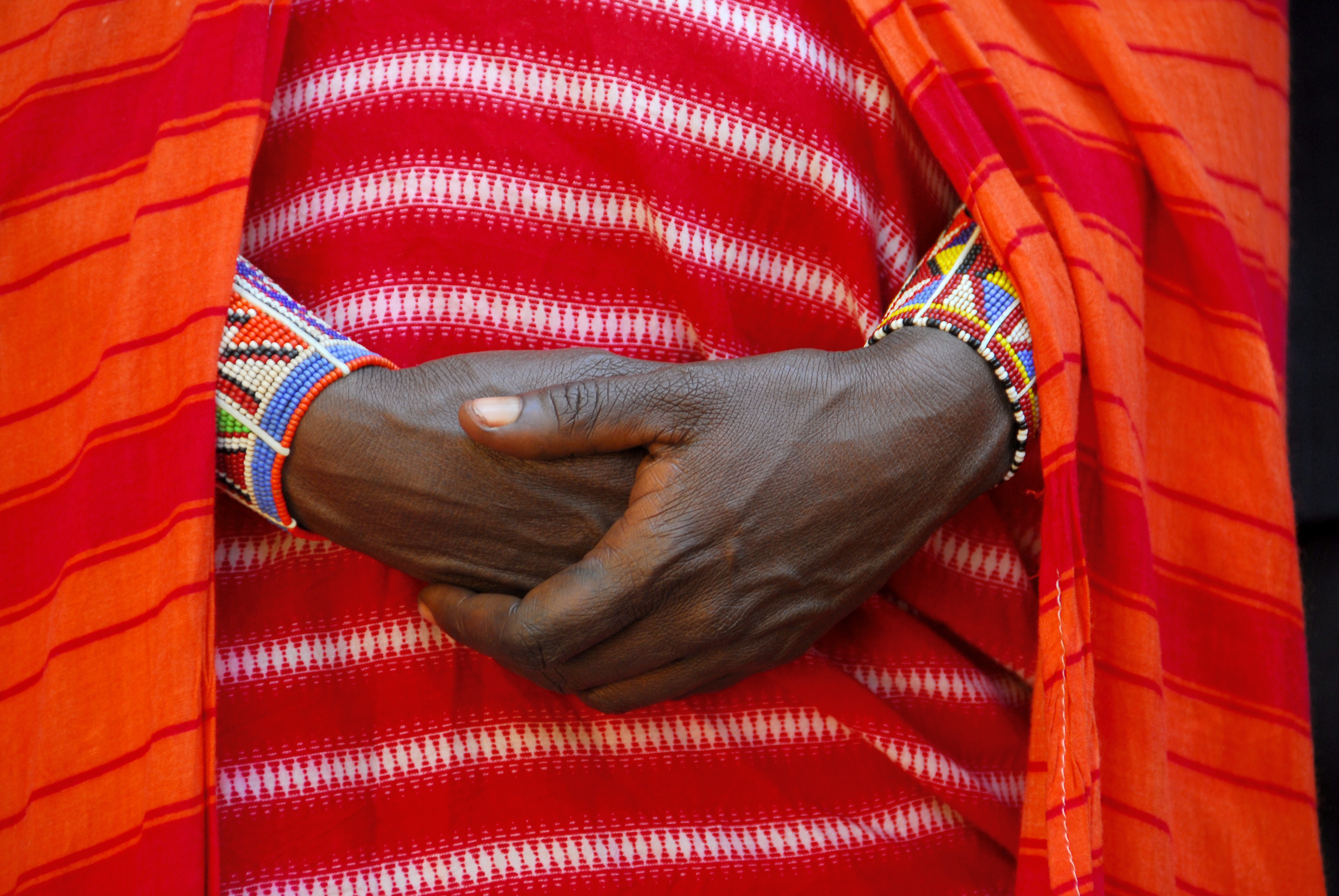 Close-up photo of a woman's hands clasped in front of her. The woman is wearing a red garment and beaded bracelets.