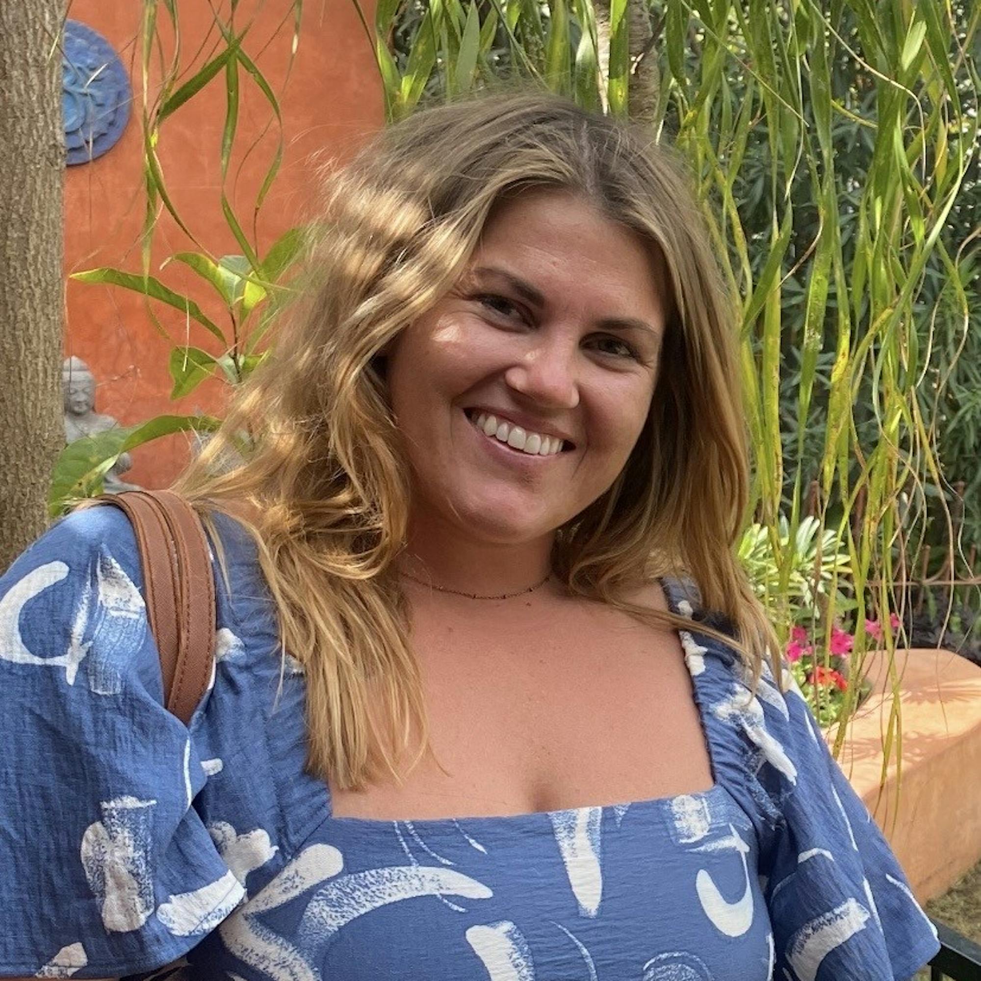 Woman smiling in a garden setting, wearing a blue patterned dress, with leafy plants and a terracotta wall in the background.