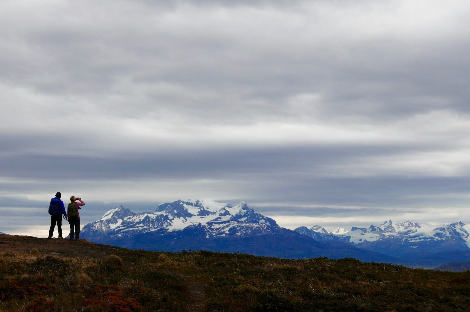 Two hikers stand on a windswept ridge in Patagonia, looking toward a broad line of snow-capped mountains.