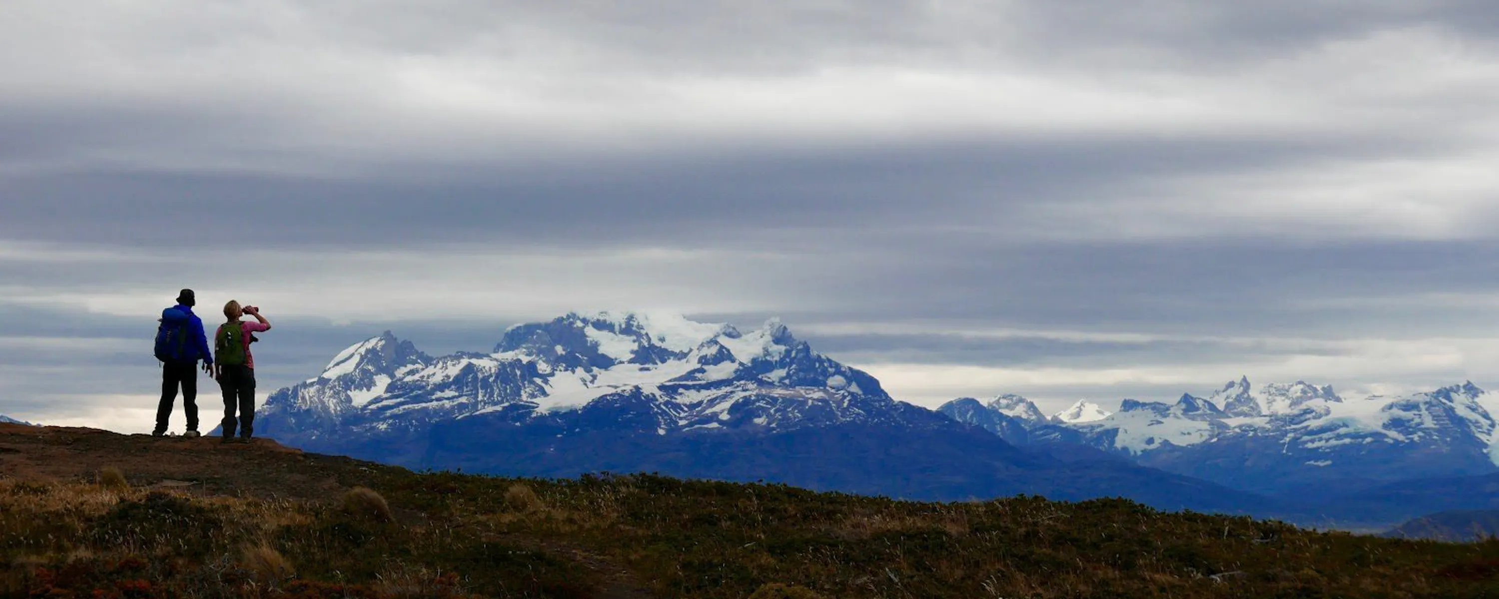 Two hikers stand on a windswept ridge in Patagonia, looking toward a broad line of snow-capped mountains.