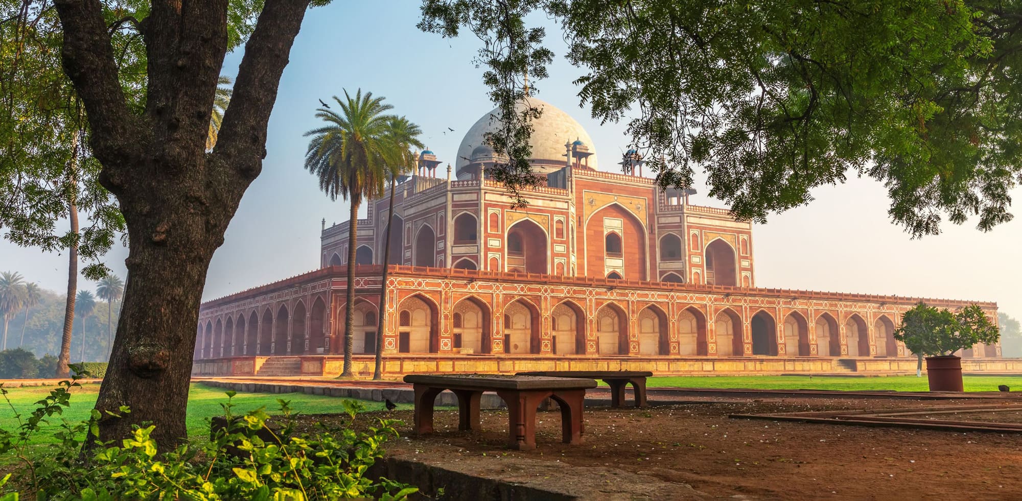Humayun's Tomb rises beyond a green lawn in Delhi, with red sandstone arcades and domes under a pale sky.