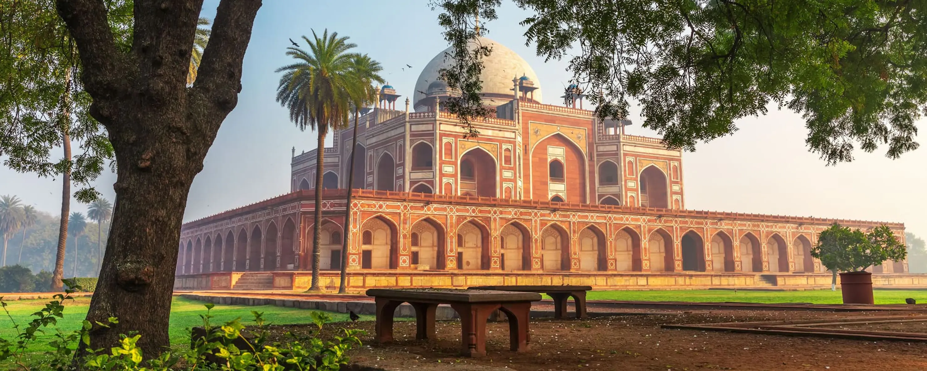 Humayun's Tomb rises beyond a green lawn in Delhi, with red sandstone arcades and domes under a pale sky.