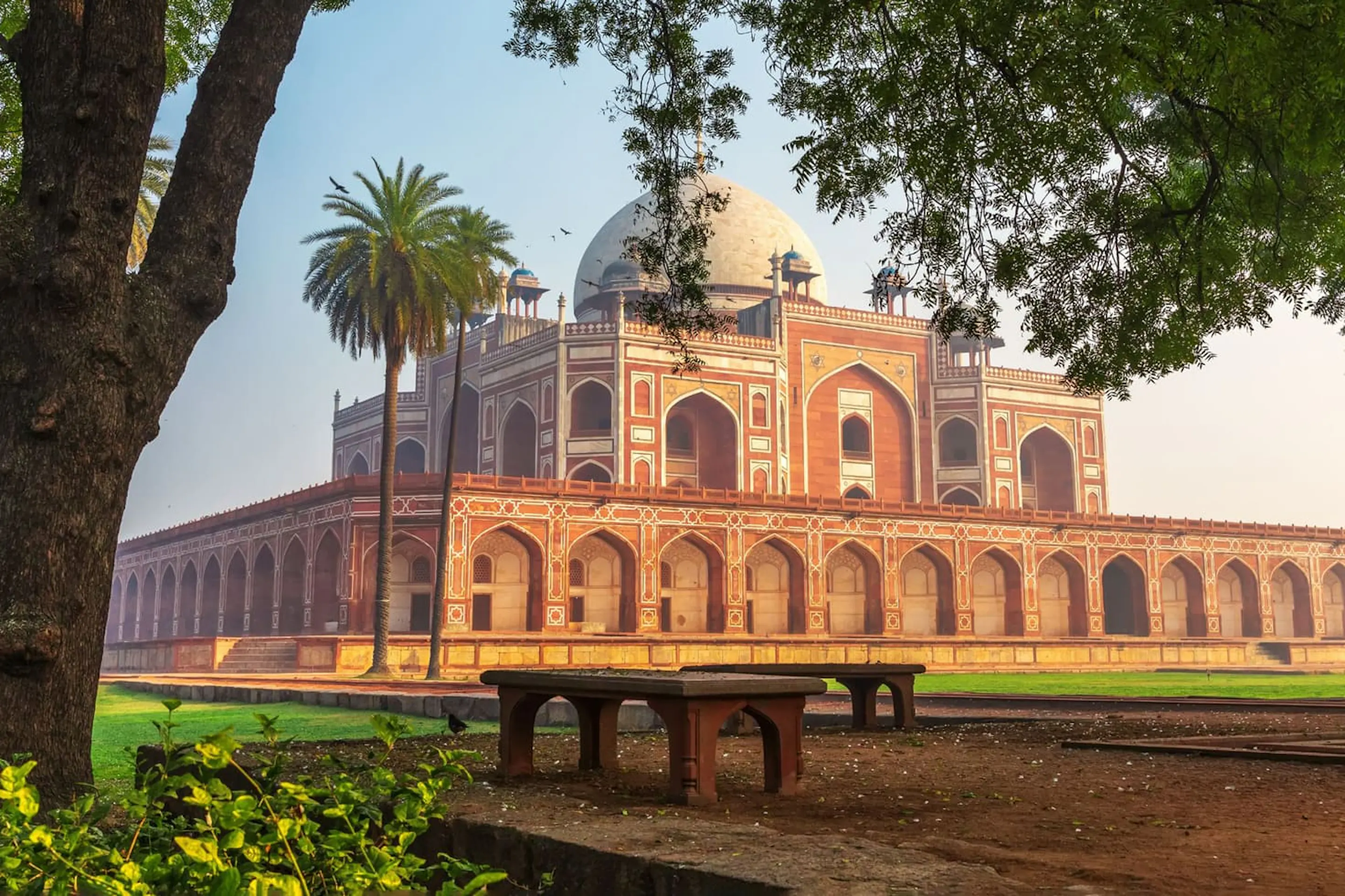 Humayun's Tomb rises beyond a green lawn in Delhi, with red sandstone arcades and domes under a pale sky.