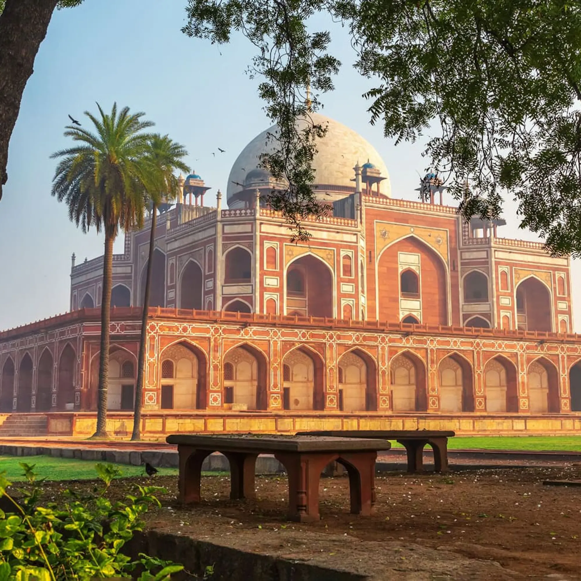 Humayun's Tomb rises beyond a green lawn in Delhi, with red sandstone arcades and domes under a pale sky.