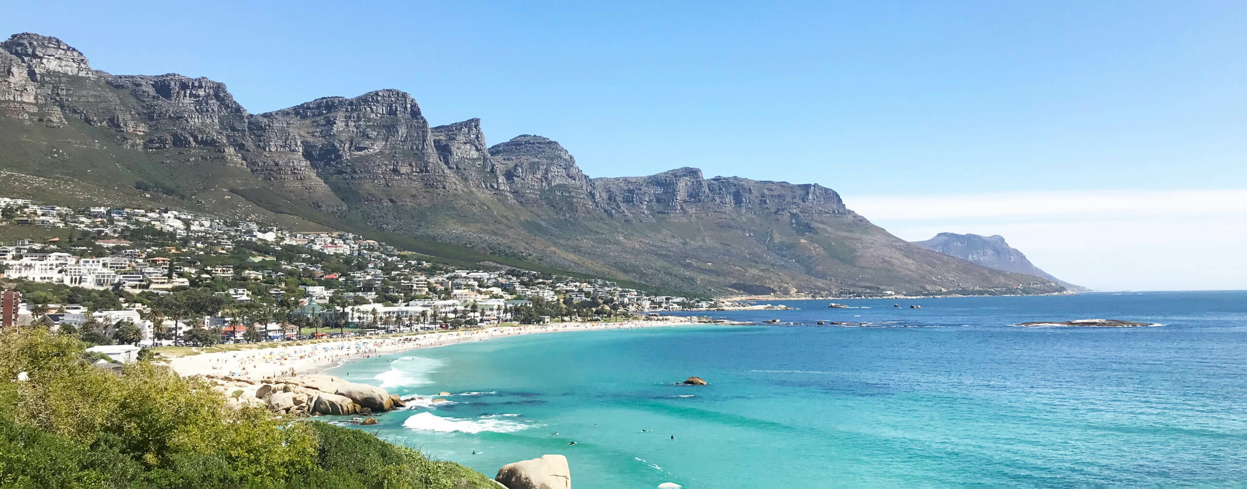 Camps Bay beach with the Twelve Apostles beyond against the backdrop of Cape Town beneath clear skies.