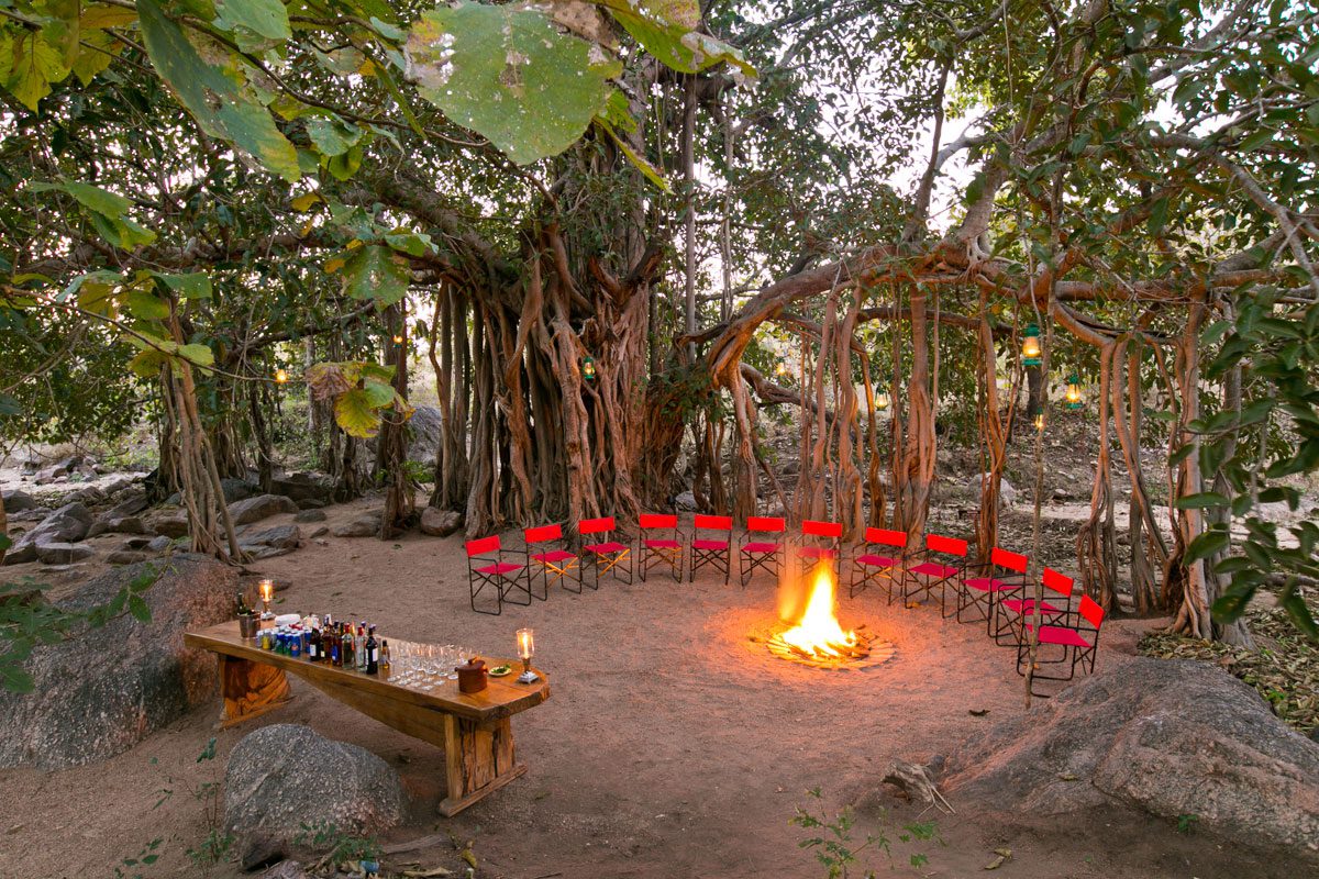 Red chairs circle a glowing fire pit at Jamtara Wilderness Camp, framed by forest and lantern light in Pench.