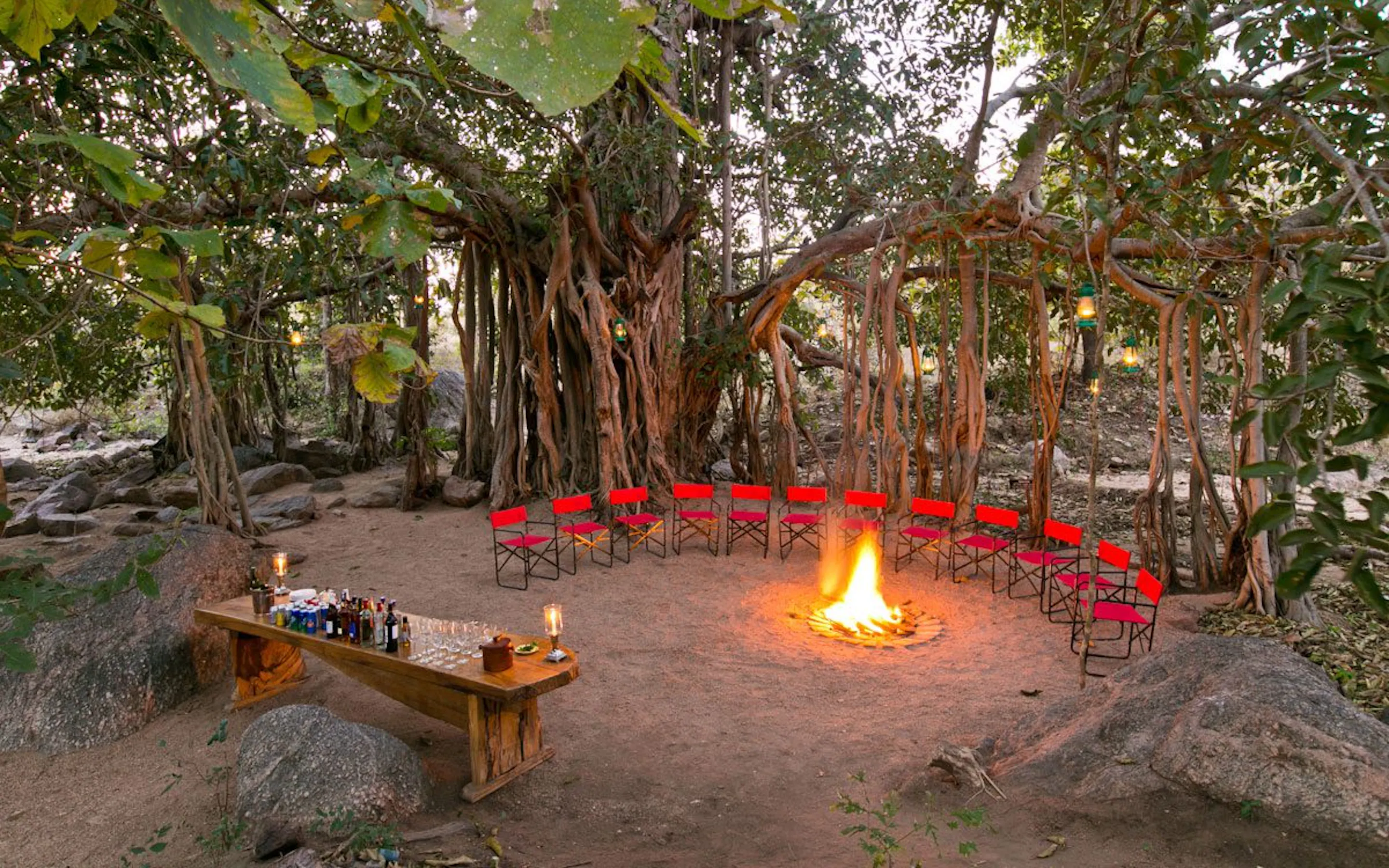 Red chairs circle a glowing fire pit at Jamtara Wilderness Camp, framed by forest and lantern light in Pench.