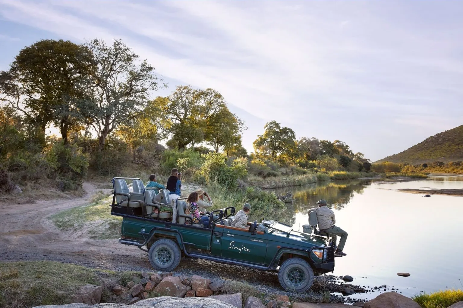 A family watches animals at the edges of a river, from an open safari vehicle in Sabi Sand