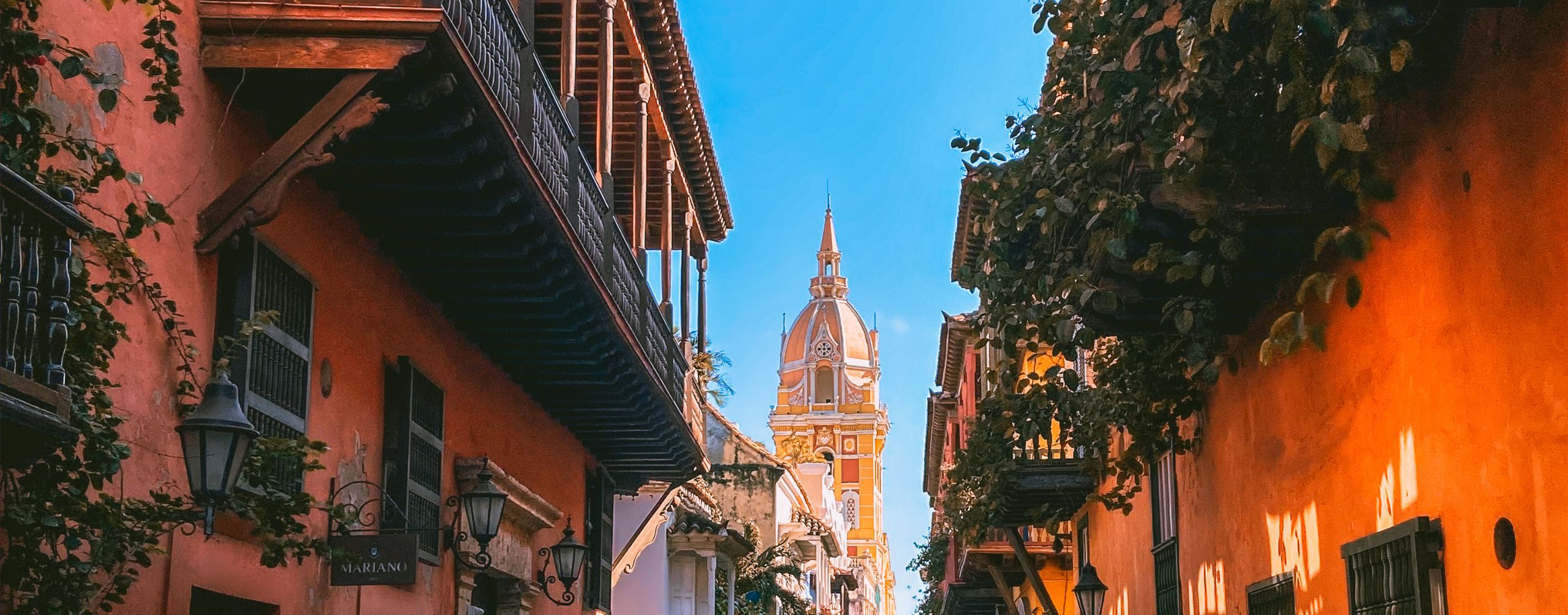 A colonial street glows with warm facades and a church tower in Cartagena, framed by balconies and hanging lights.