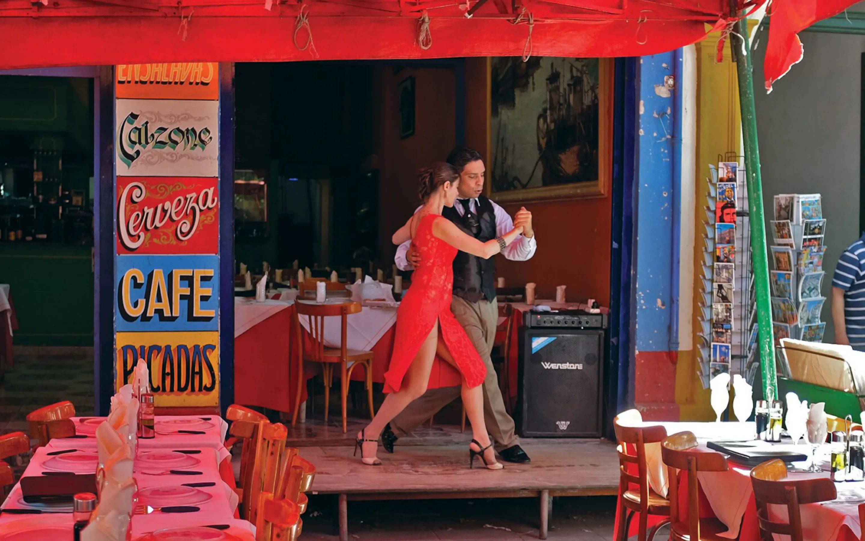 A couple dances in a bright red cafe doorway in La Boca, framed by colorful walls, striped awnings, and empty tables.