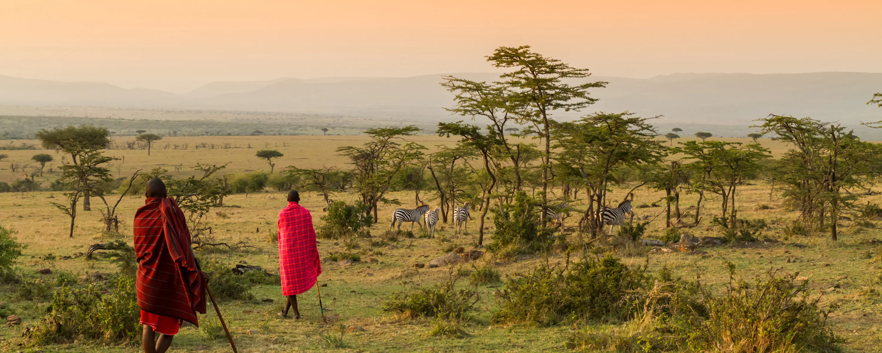 Two Maasai stand above Kenya's open plains, looking across golden grass under a dramatic sky near the Mara.