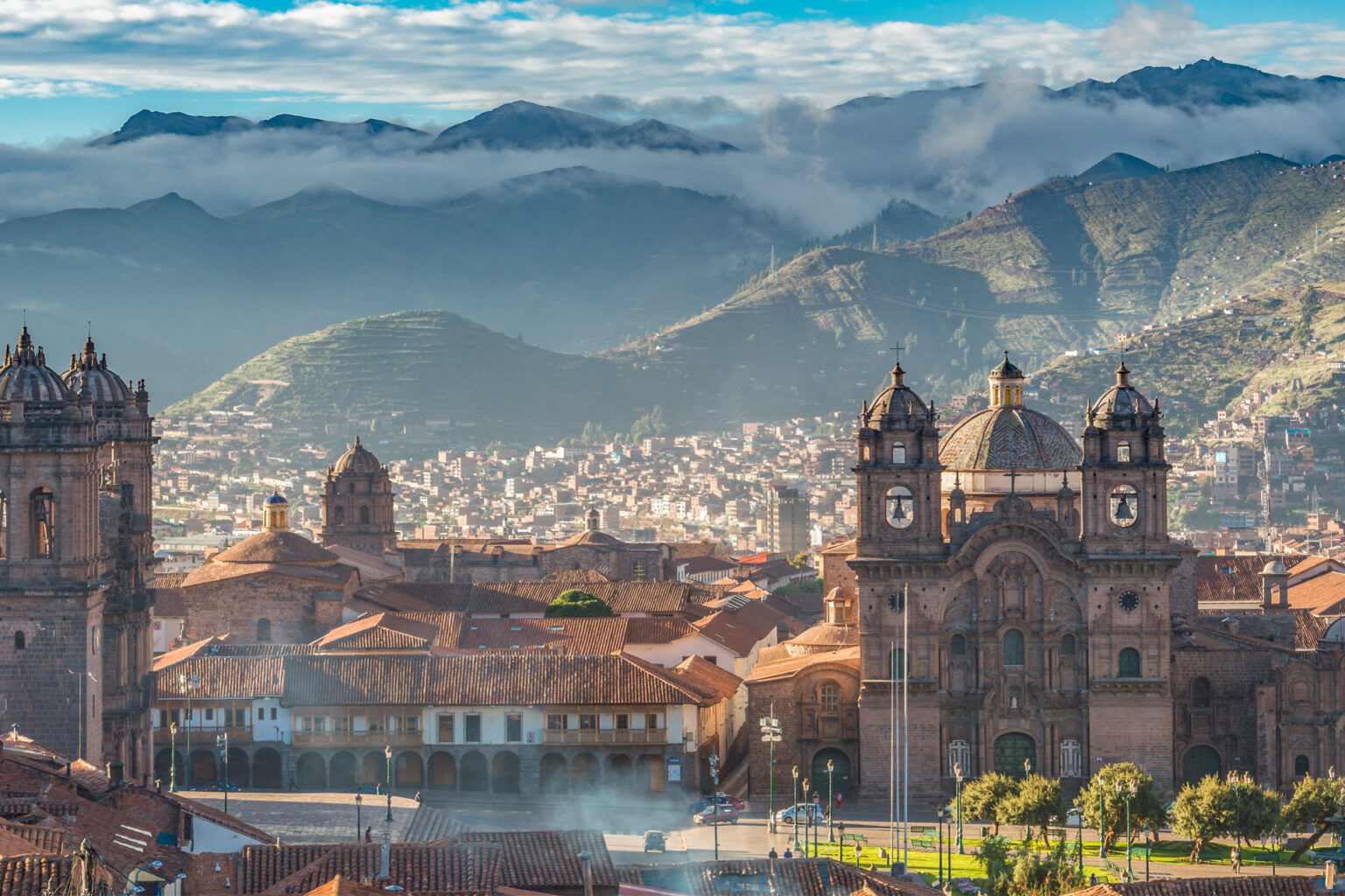 Cusco rooftops and cathedral towers rise beneath layered mountains in Peru, with tiled buildings filling the foreground.