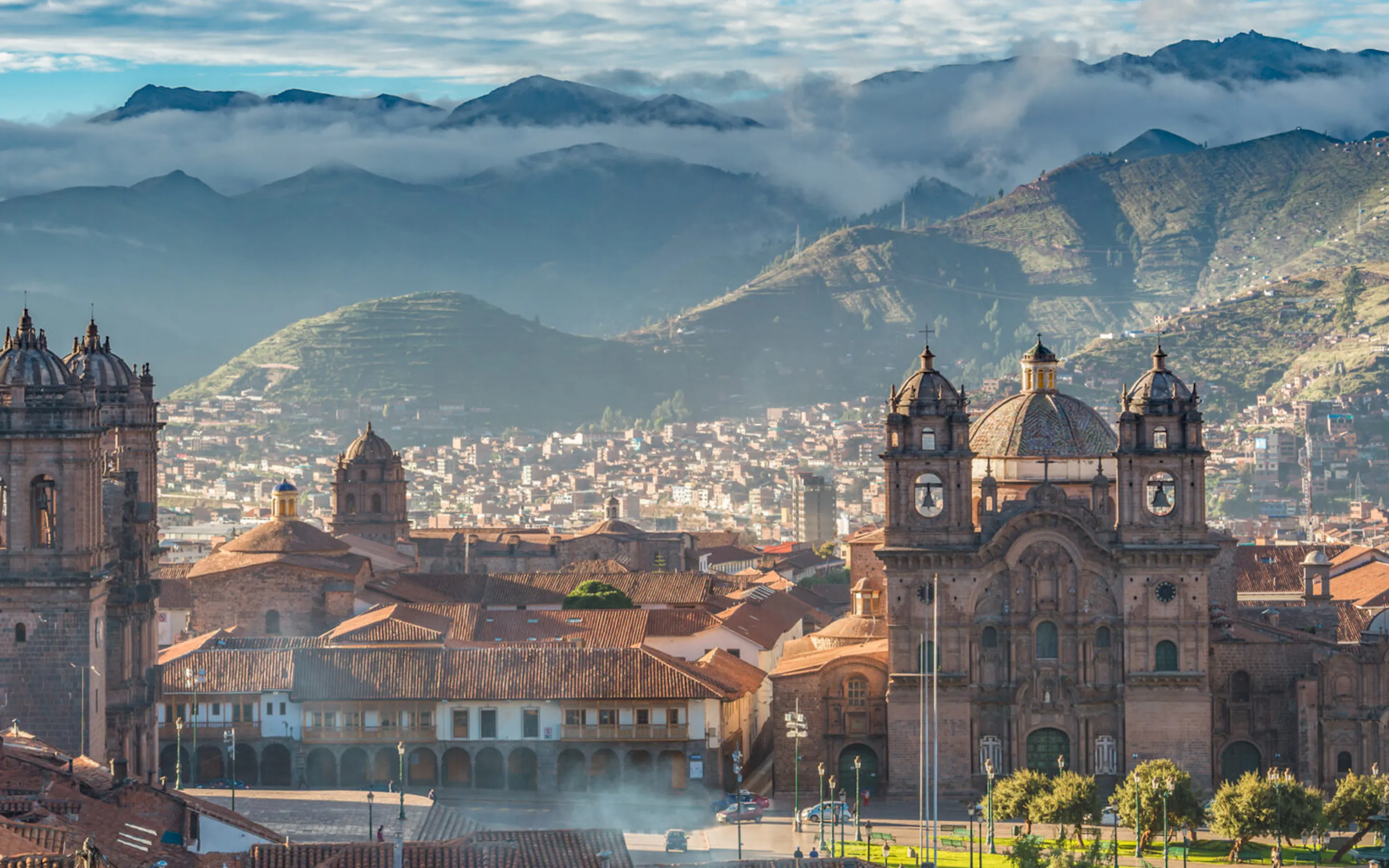 Cusco rooftops and cathedral towers rise beneath layered mountains in Peru, with tiled buildings filling the foreground.