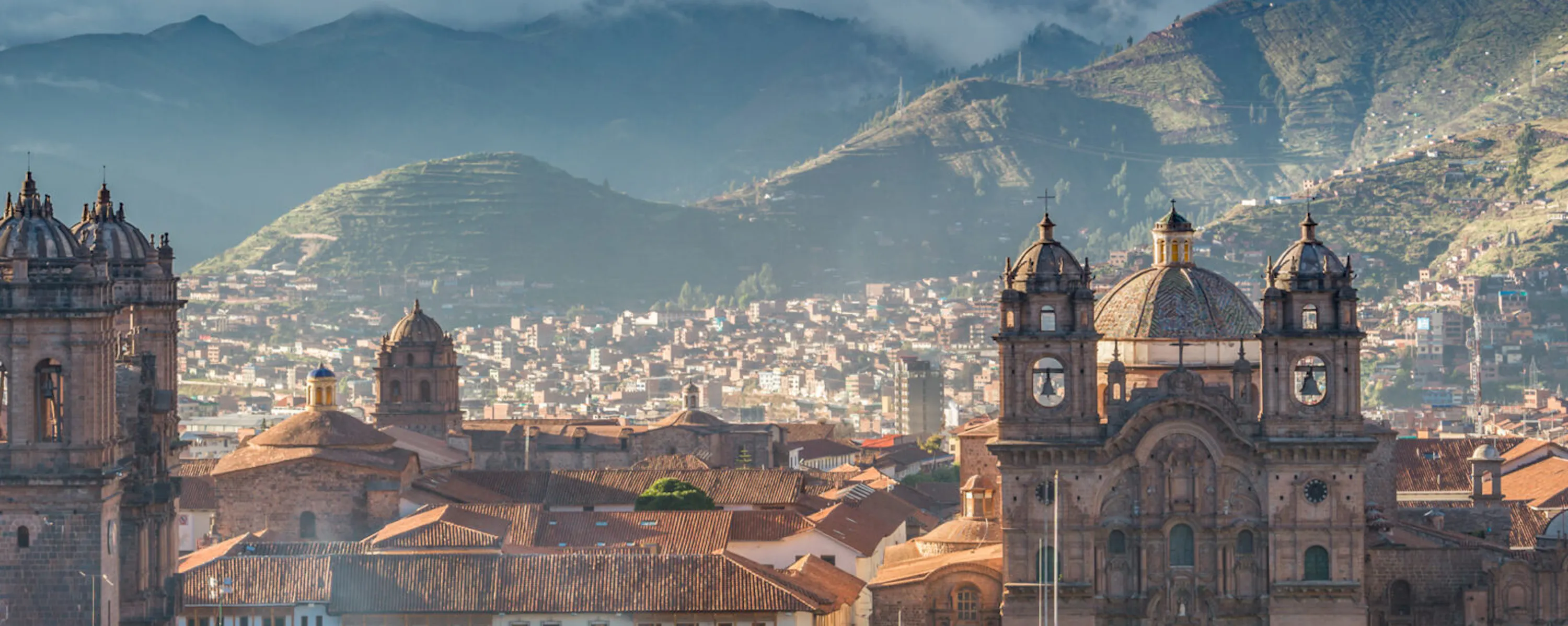 Cusco rooftops and cathedral towers rise beneath layered mountains in Peru, with tiled buildings filling the foreground.