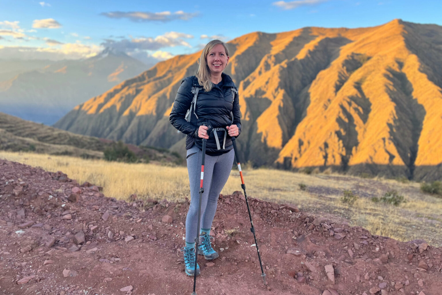 A hiker stands above Peru's Sacred Valley, with striped mountains and long shadows stretching across the slope.