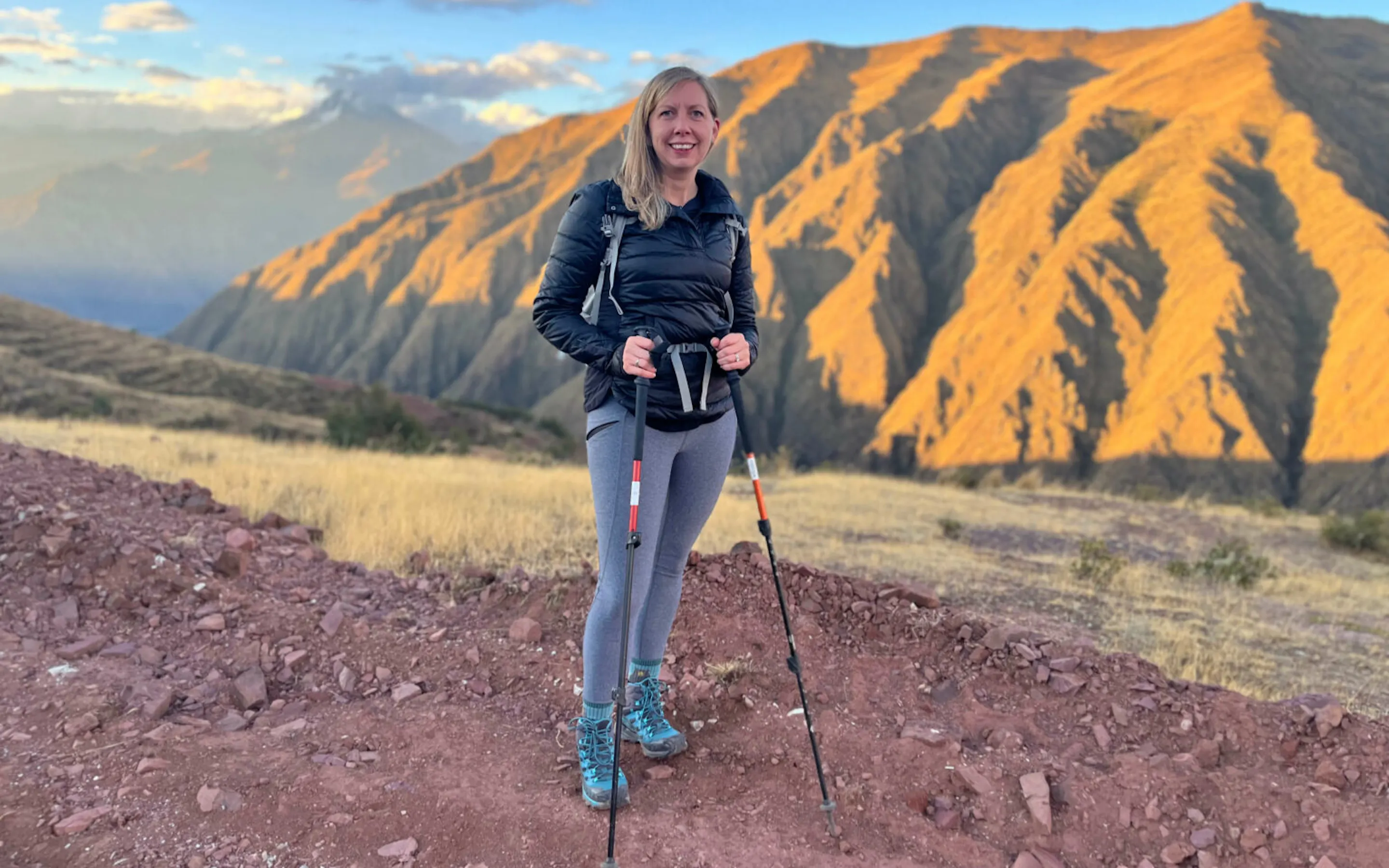 A hiker stands above Peru's Sacred Valley, with striped mountains and long shadows stretching across the slope.