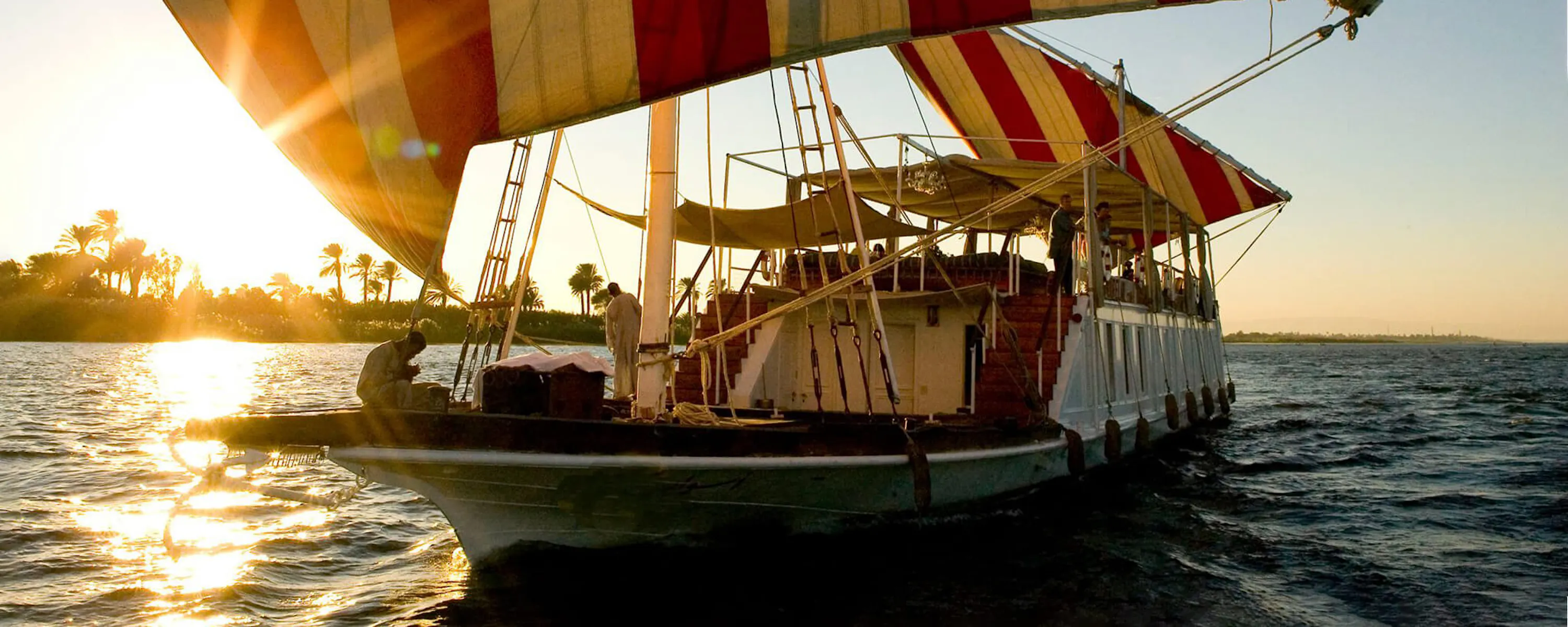 A striped sailboat glides across the Nile at Nour El Nil in Egypt, with calm water and a fading sky around it.