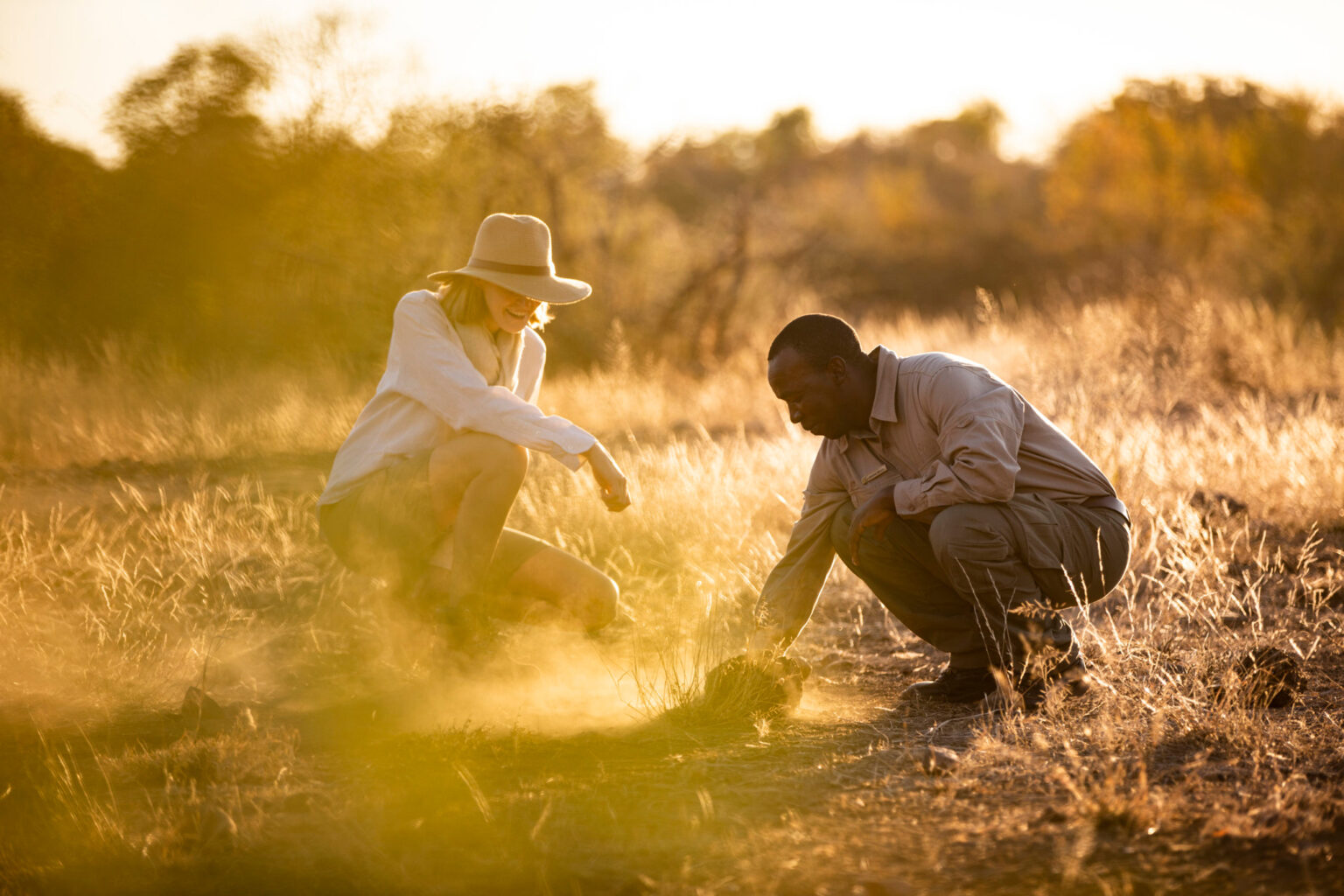 A guide and guest crouching in golden grass at sunset against the backdrop of South Africa's Greater Kruger.