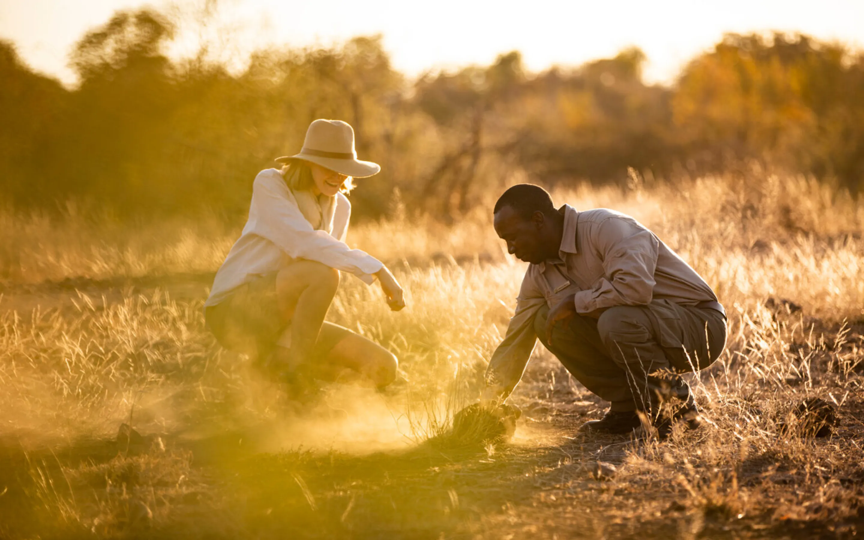 A guide and guest crouching in golden grass at sunset against the backdrop of South Africa's Greater Kruger.