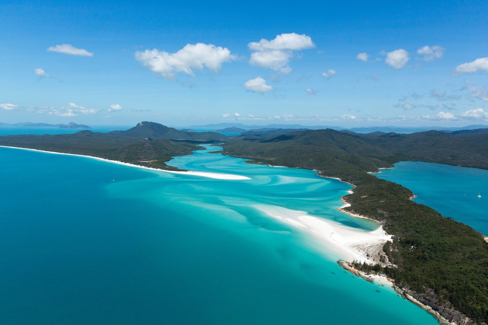 A long sandbar curls through bright blue water in the Whitsundays, with forested islands fading into the distance.