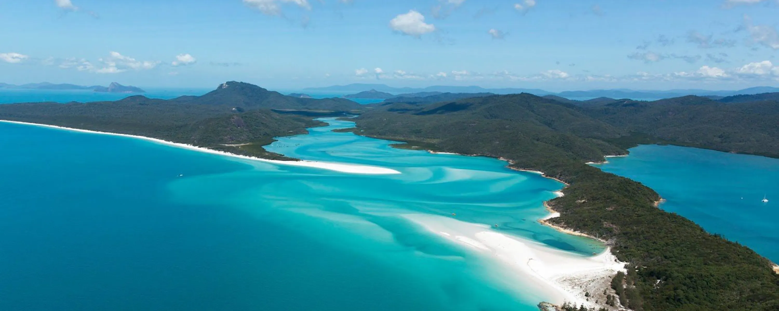 A long sandbar curls through bright blue water in the Whitsundays, with forested islands fading into the distance.
