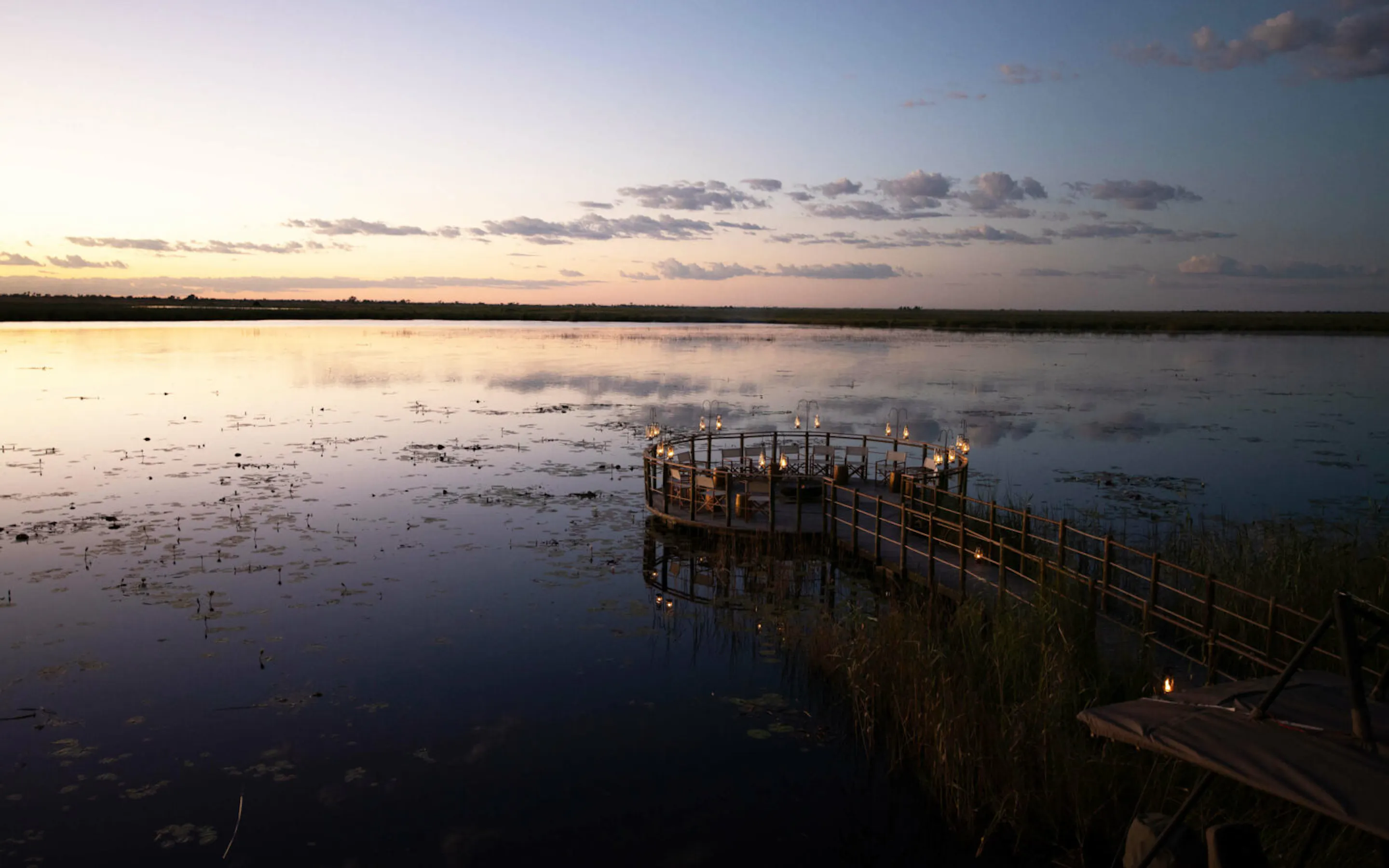 A candlelit deck floating above calm delta waters at dusk against the backdrop of Botswana's Okavango Delta.