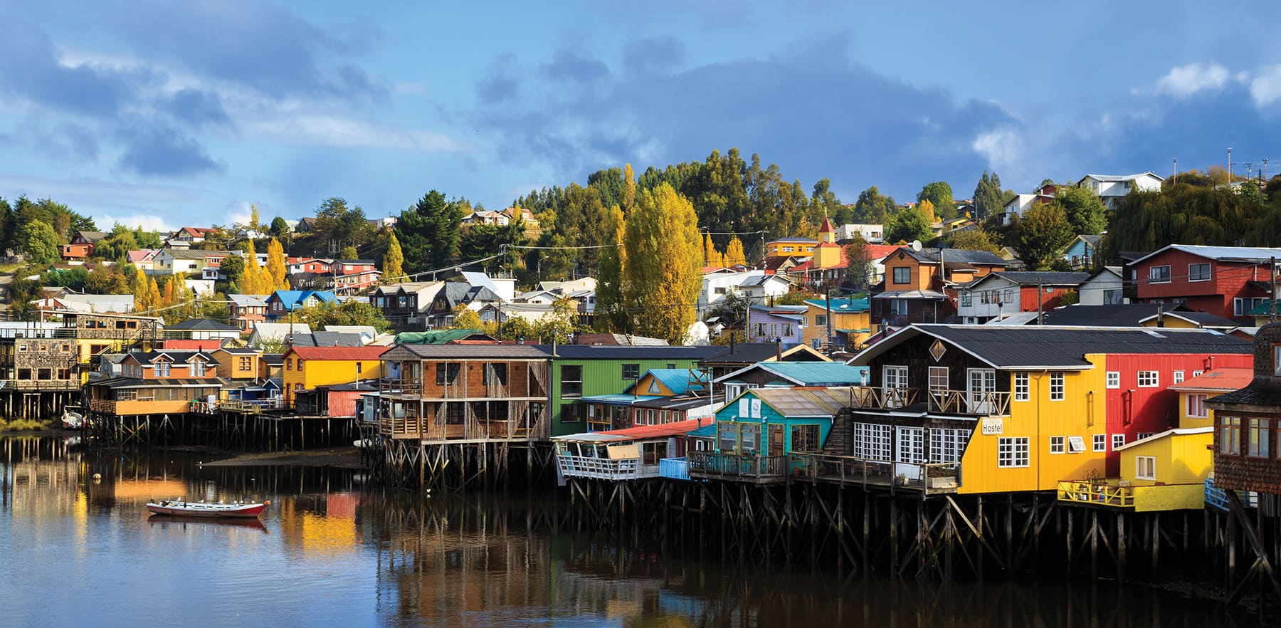 Colorful stilt houses lining calm waterfront reflections in Chile's Chiloe Island, under expansive skies.