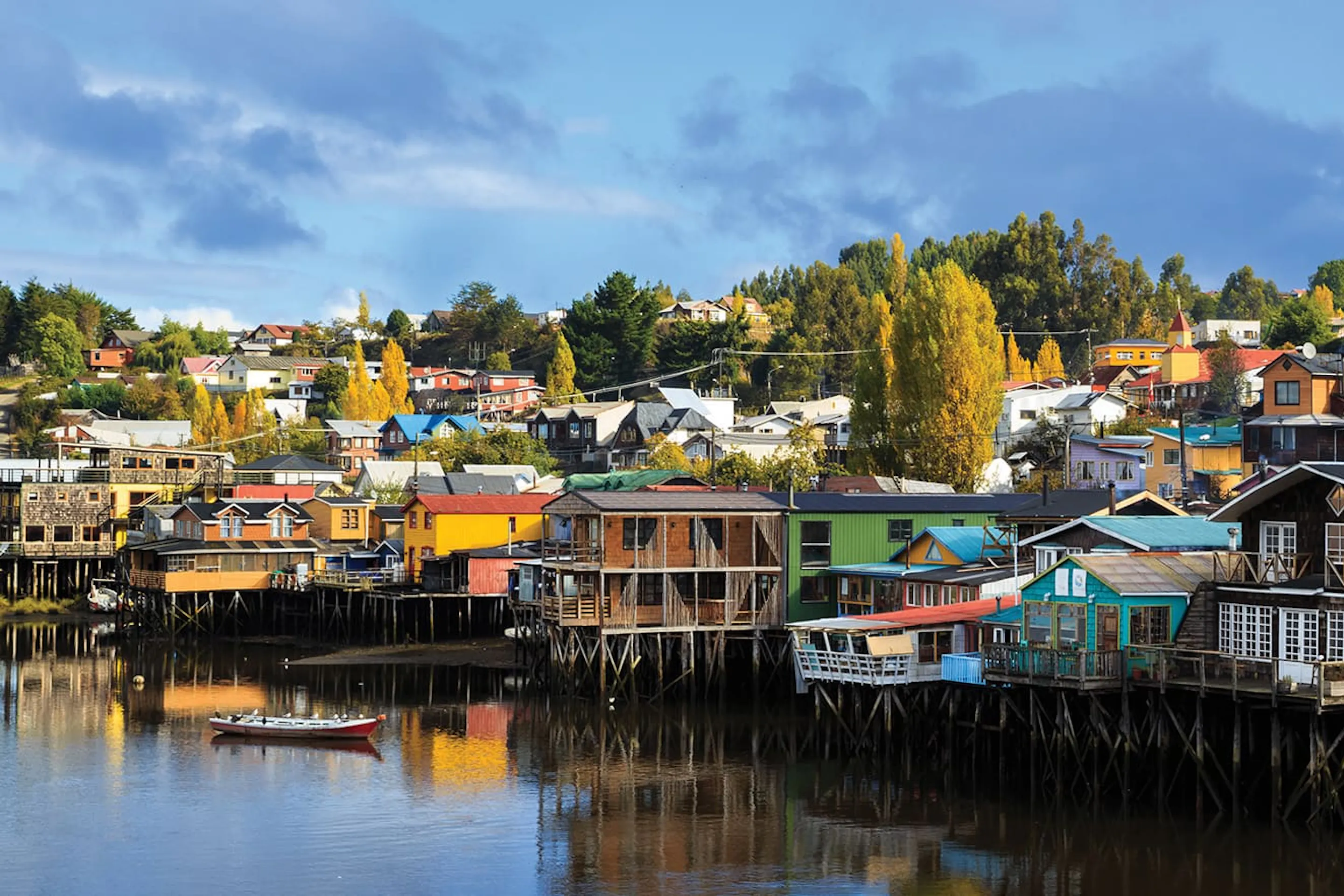 Colorful stilt houses lining calm waterfront reflections in Chile's Chiloe Island, under expansive skies.