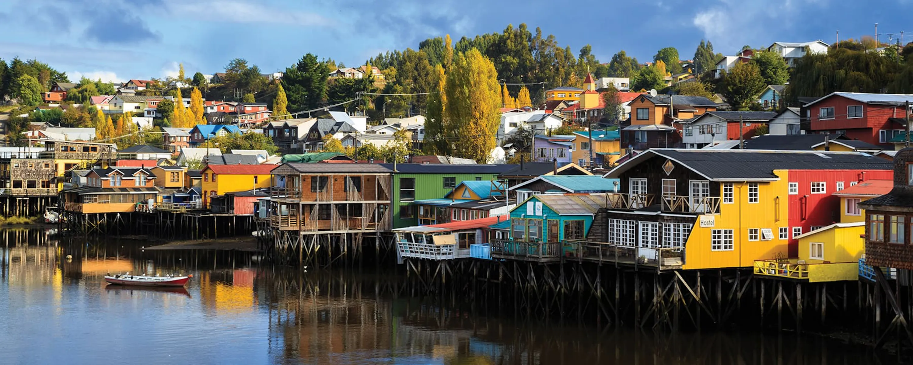 Colorful stilt houses lining calm waterfront reflections in Chile's Chiloe Island, under expansive skies.