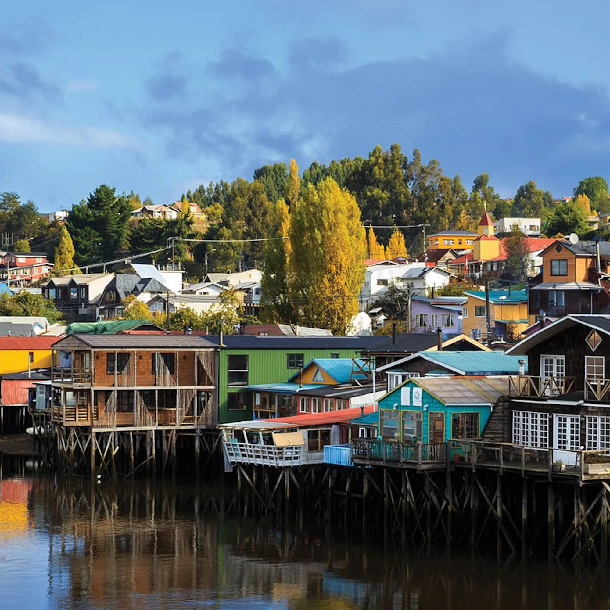 Colorful stilt houses lining calm waterfront reflections in Chile's Chiloe Island, under expansive skies.