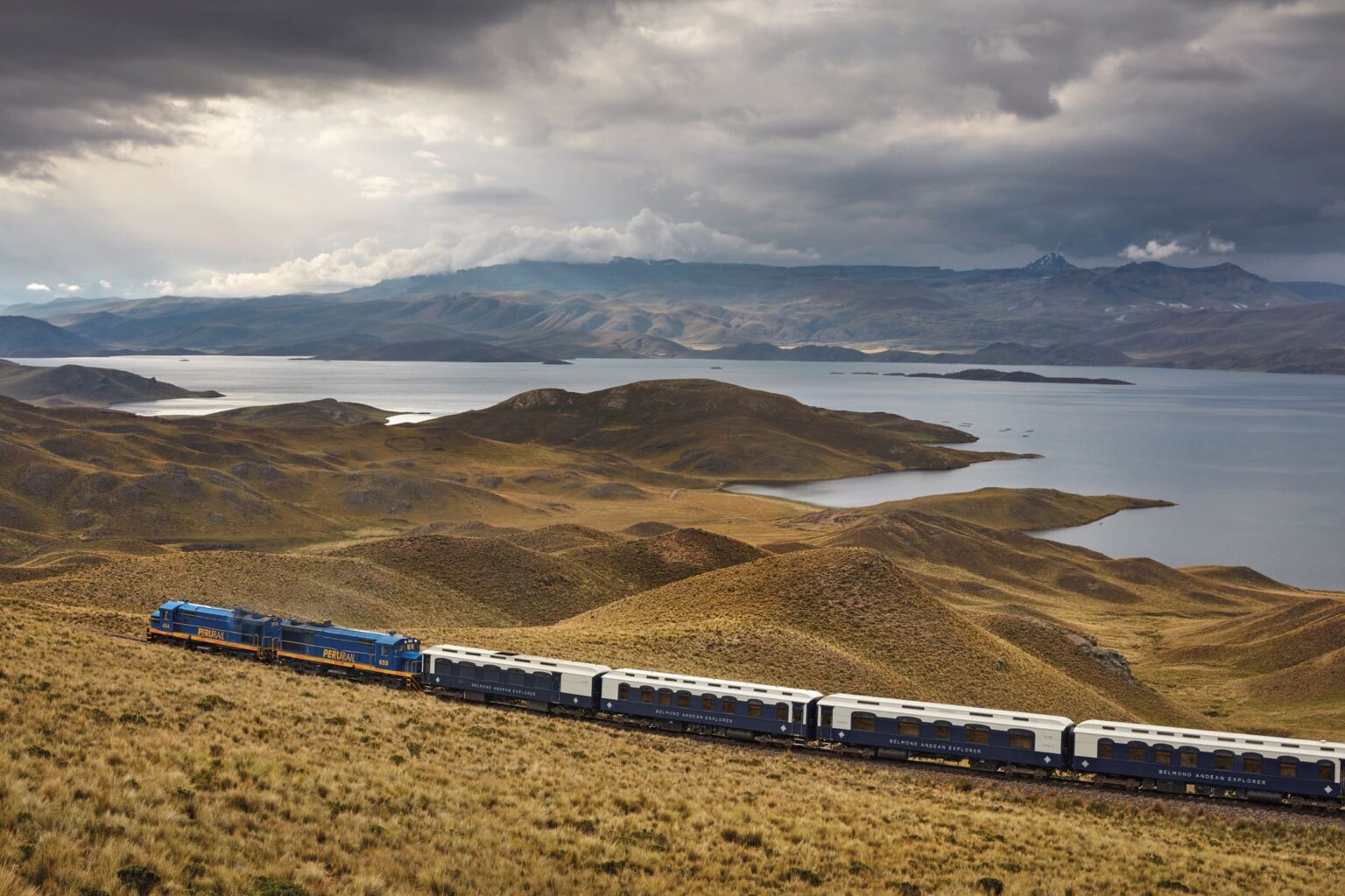 The Belmond Andean Explorer winds through Peru's high plains, with lakes and distant mountains under dramatic cloud.