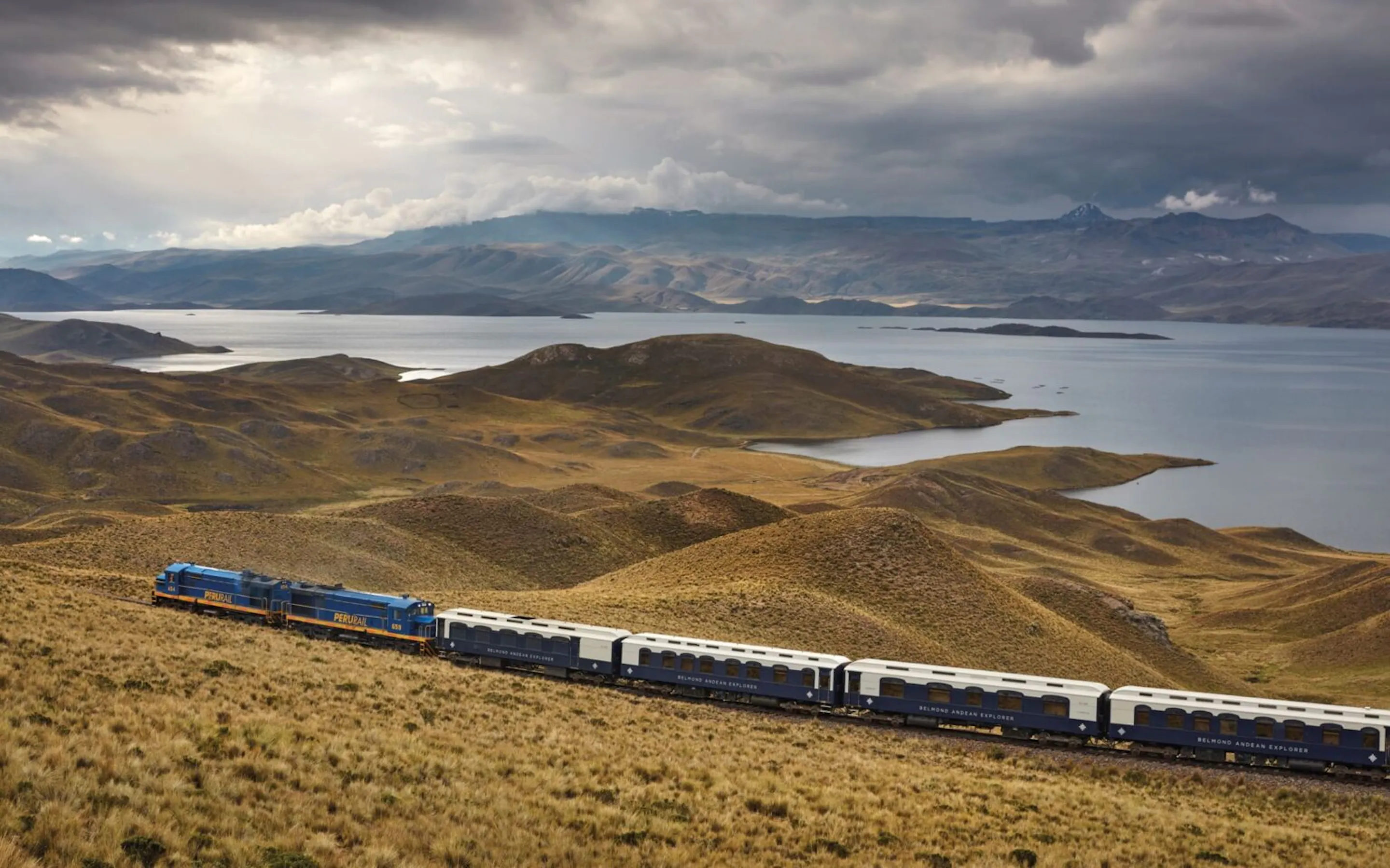 The Belmond Andean Explorer winds through Peru's high plains, with lakes and distant mountains under dramatic cloud.