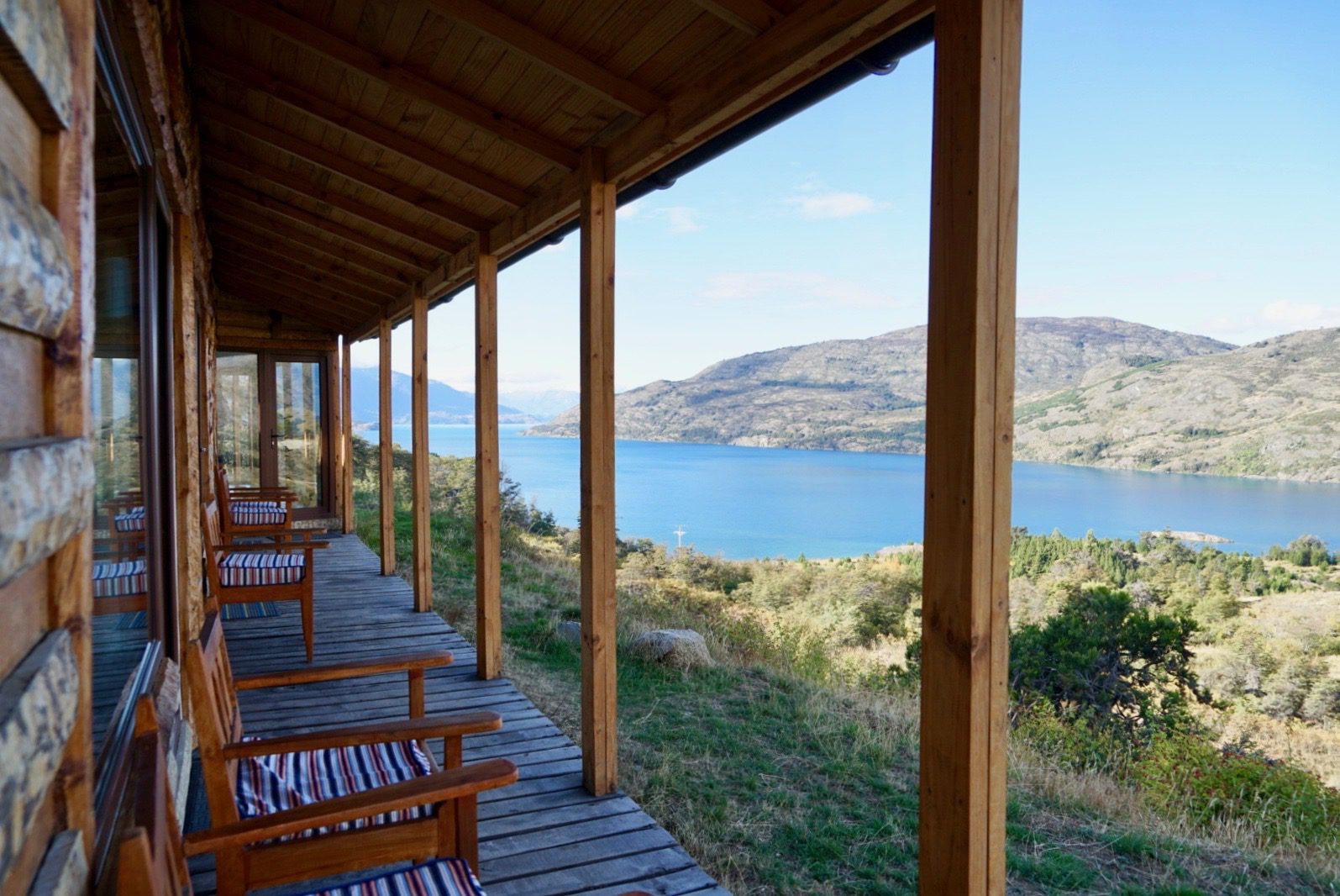 Wooden chairs line a covered porch at Mallin Colorado, overlooking Lake General Carrera and pale hills beyond.