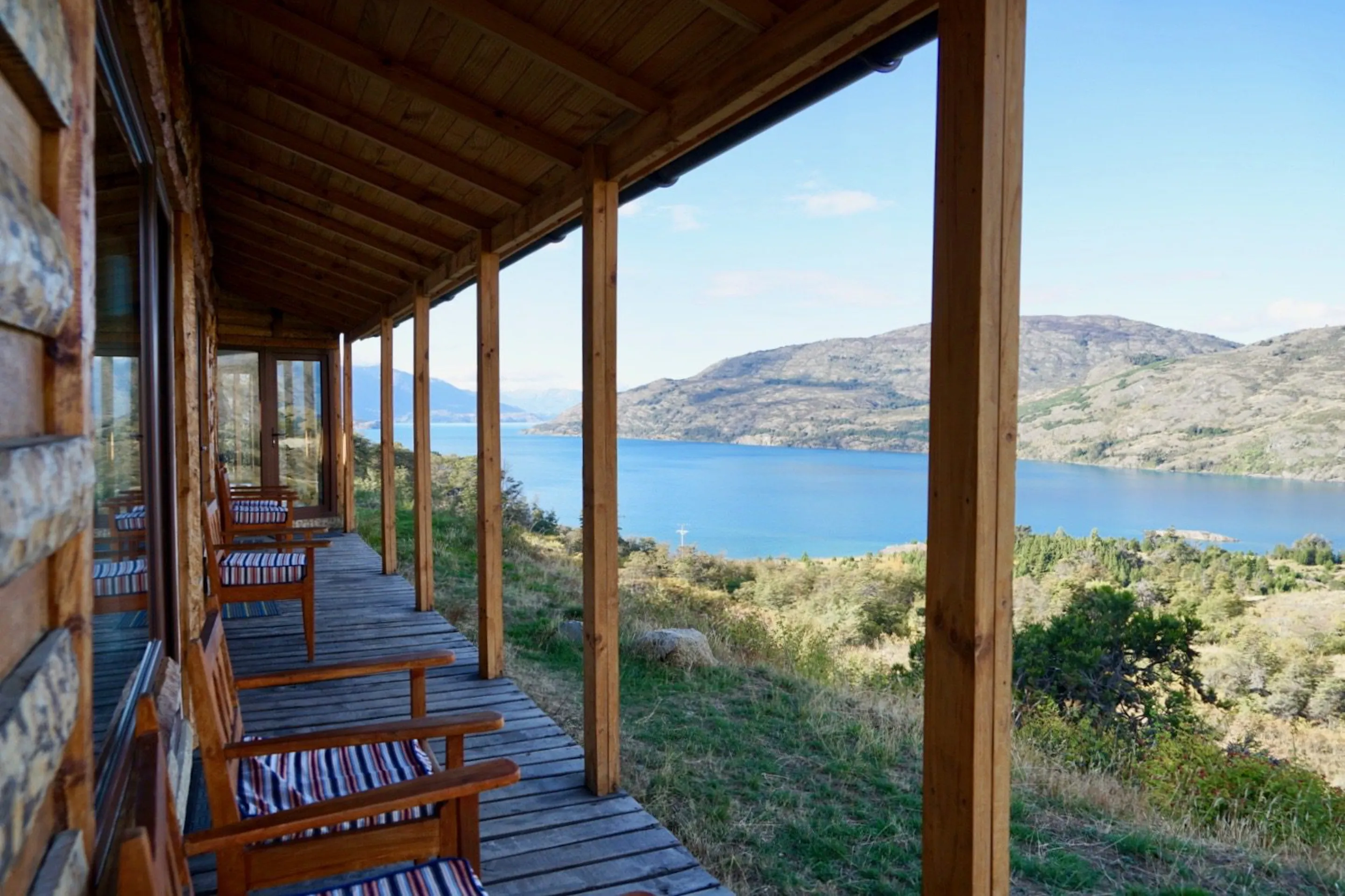 Wooden chairs line a covered porch at Mallin Colorado, overlooking Lake General Carrera and pale hills beyond.