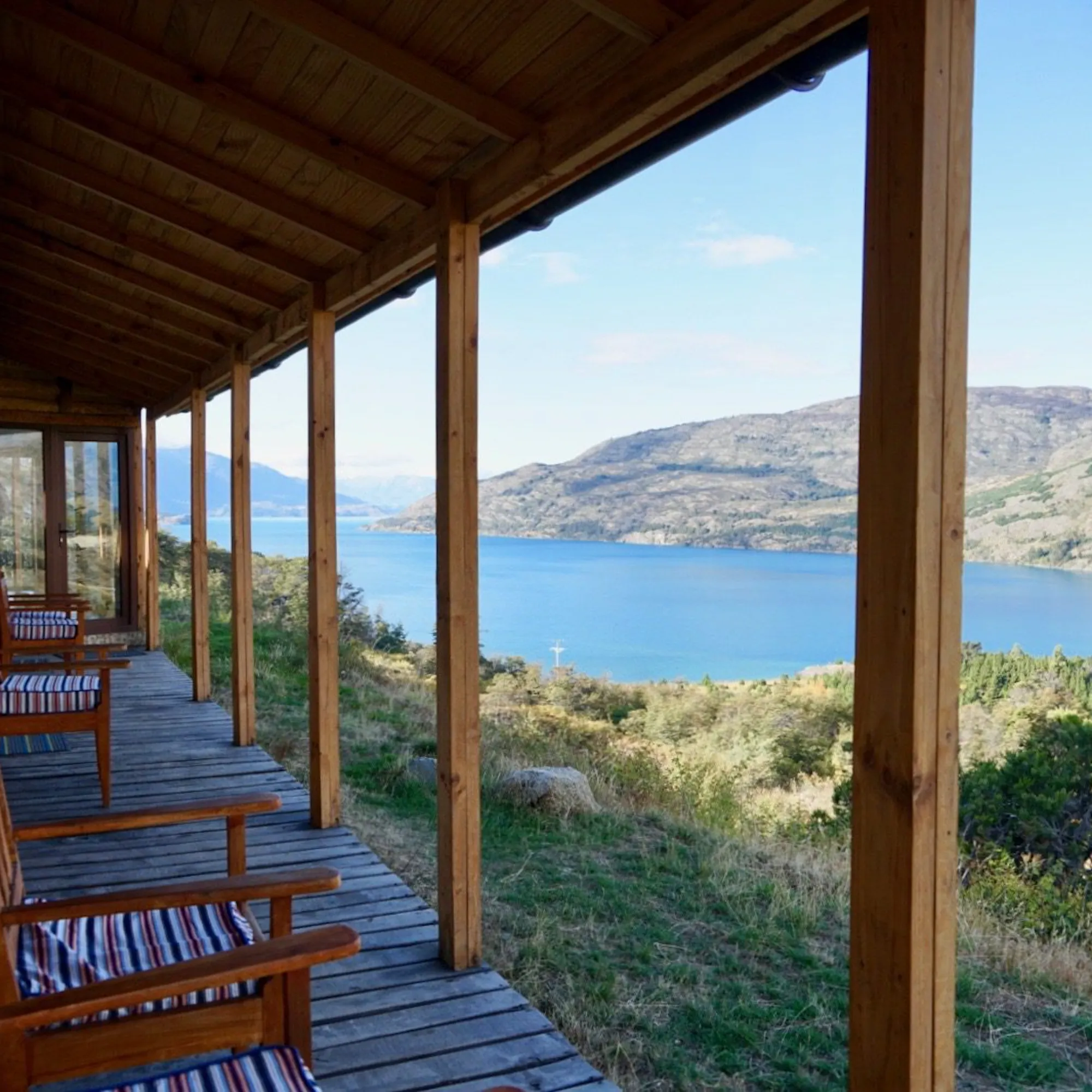 Wooden chairs line a covered porch at Mallin Colorado, overlooking Lake General Carrera and pale hills beyond.