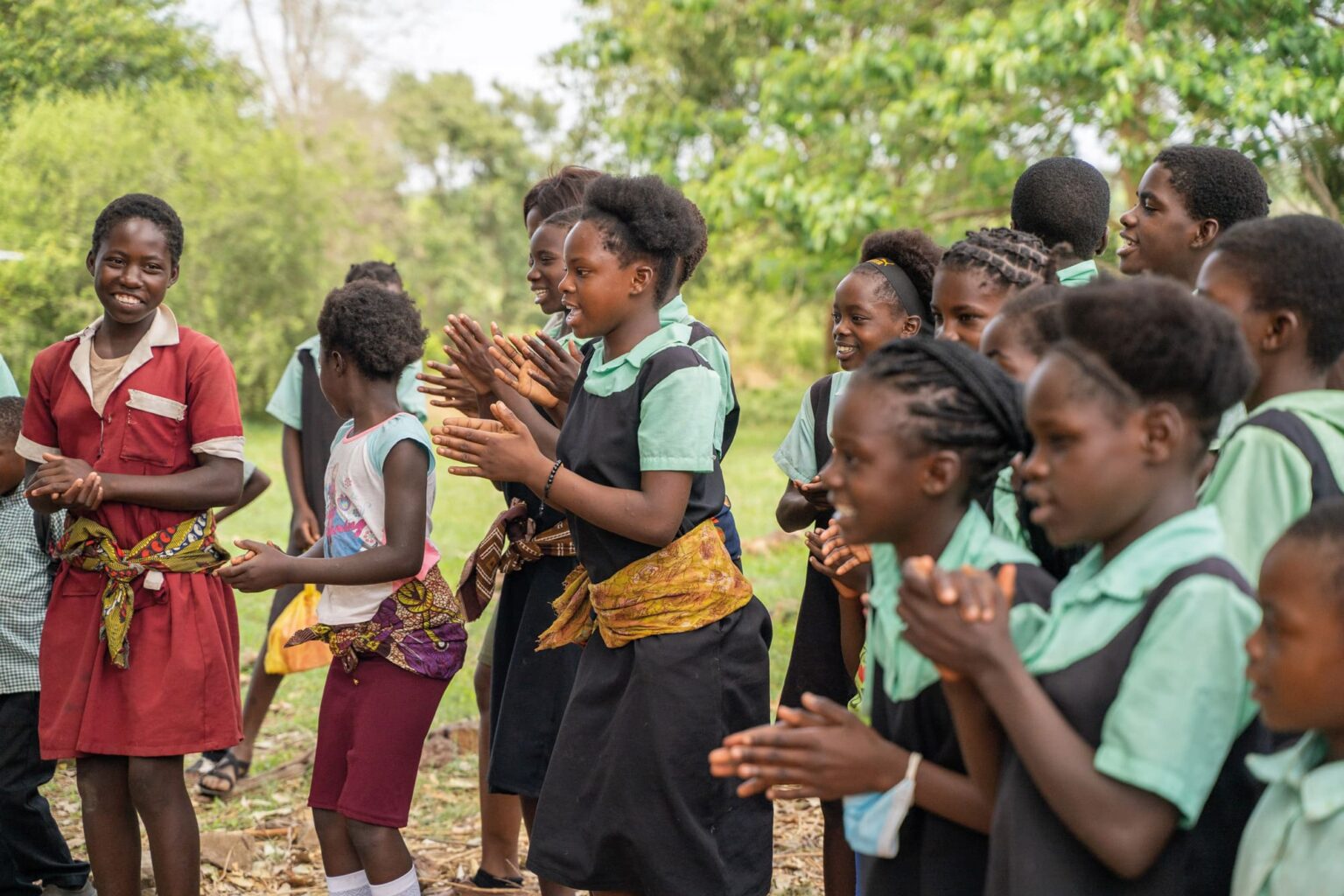 Schoolgirls in colorful uniforms clap and sing outdoors, gathered in a sunlit community scene with trees behind them.